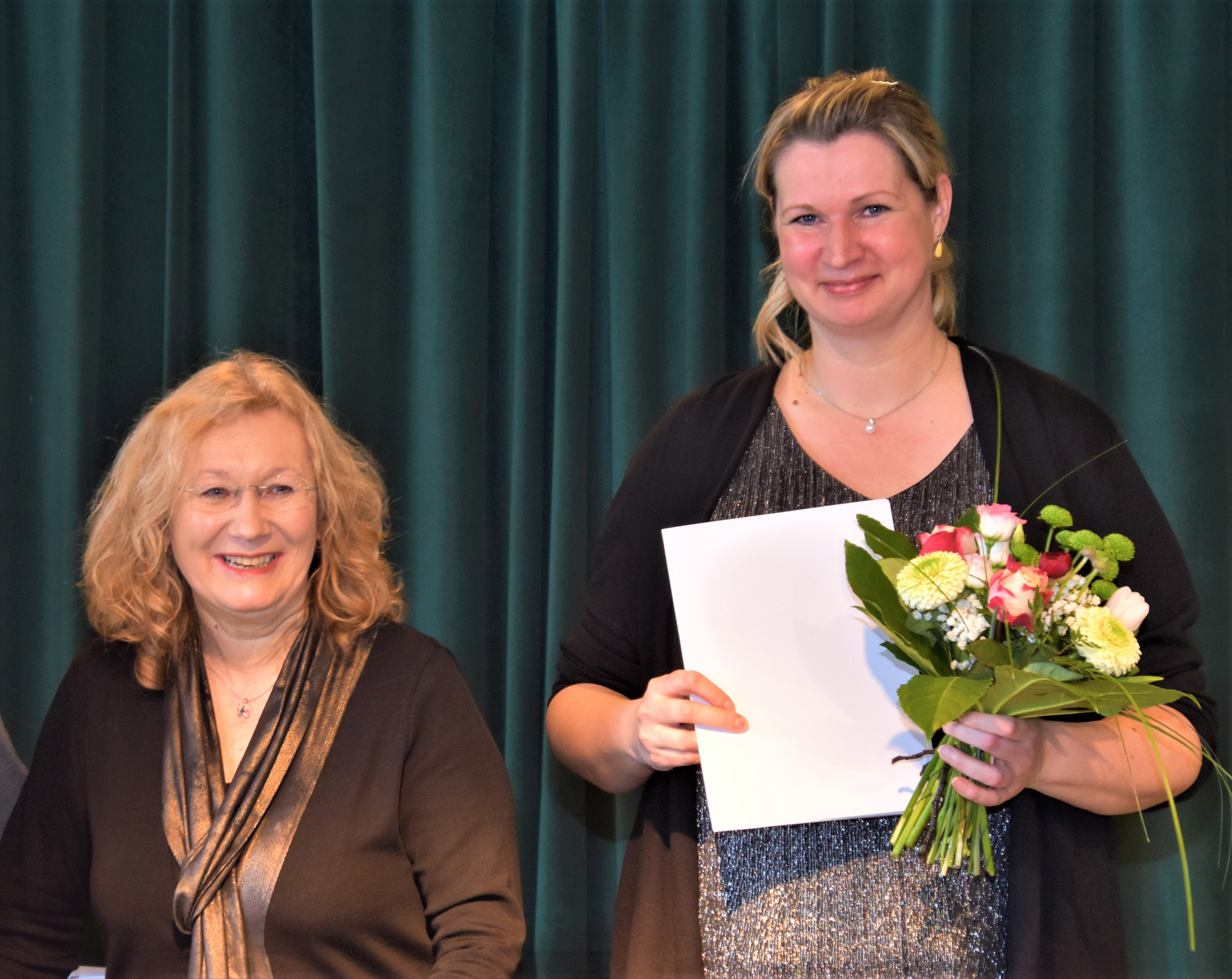 Zwei Frauen vor einem dunkelgrünen Vorhang, eine mit Zeugnis und Blumenstrauß in den Händen. Beide lächeln in die Kamera.