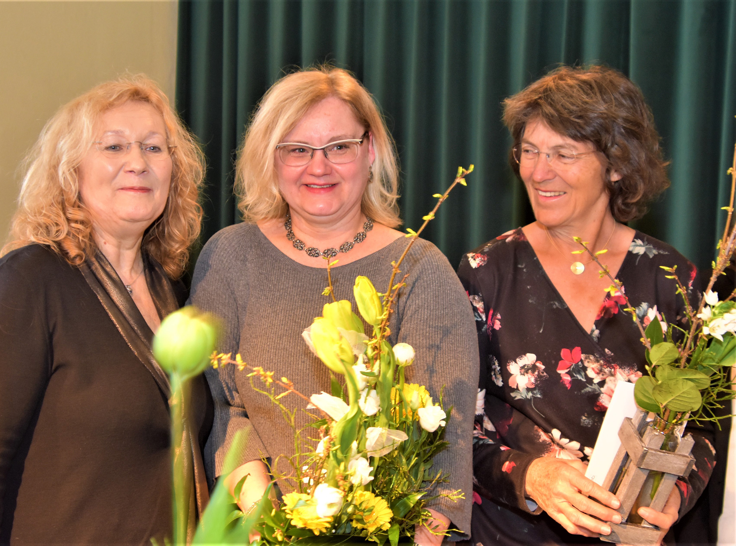 Drei Frauen vor dunkelgrünem Vorhand mit kleinen Blumengestecken in der Hand. Alle lächeln fröhlich.