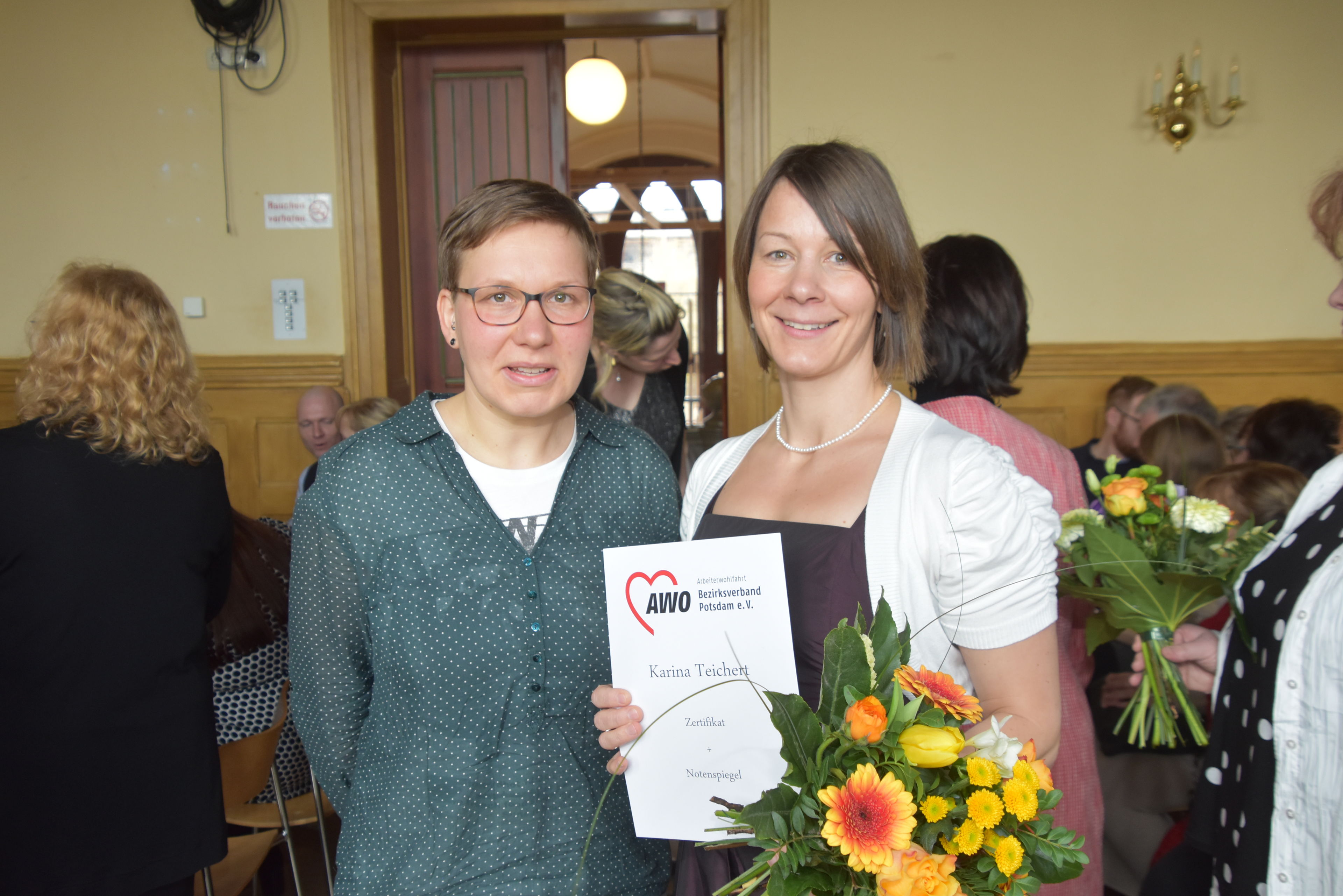 Zwei Frauen in einem Raum. Eine hält Zeugnis und Blumen in den Händen. Lächeln in die Kamera. Andere Personen im Hintergrund.