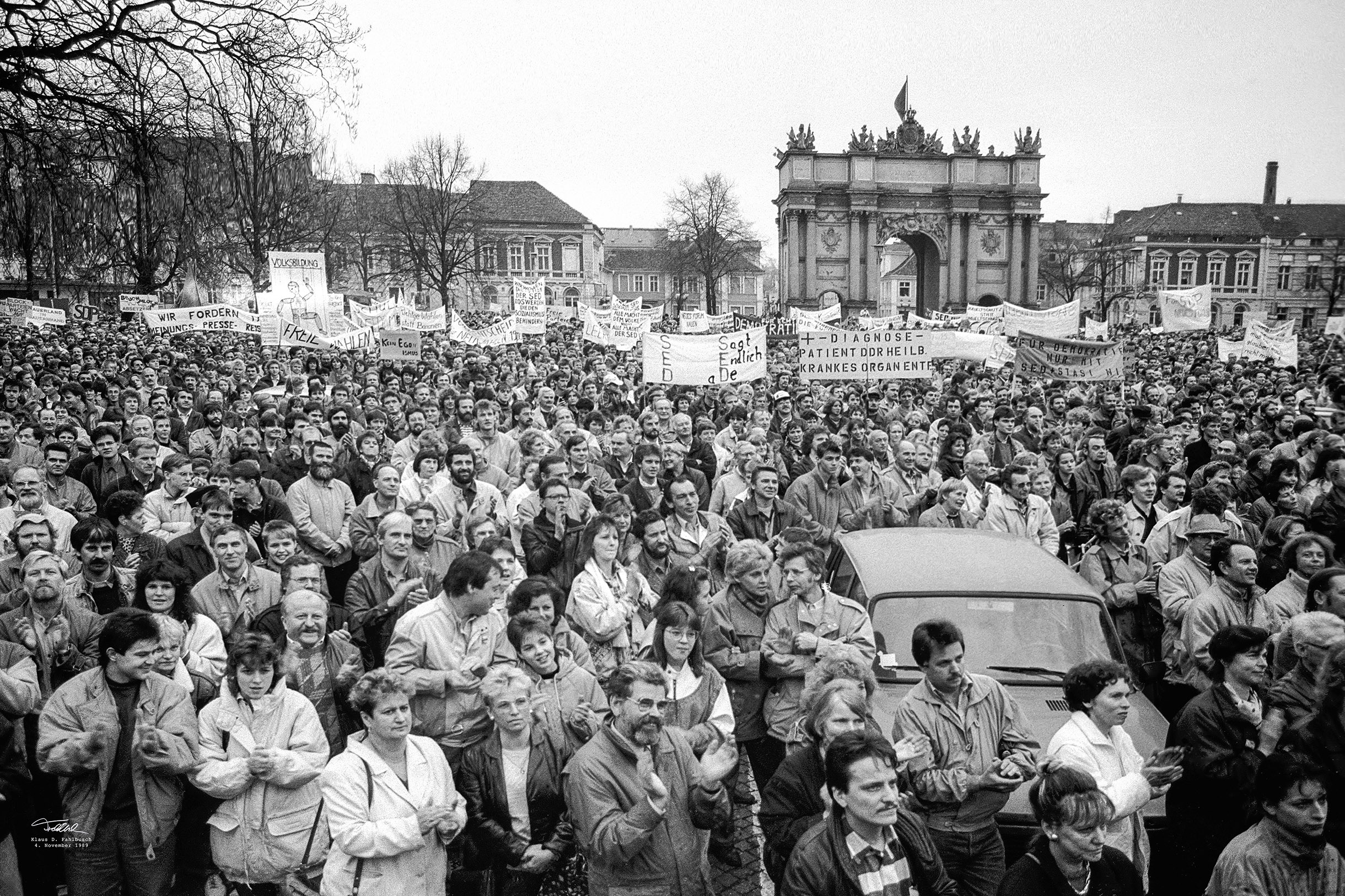 Schwarz-Weiß Foto: Vor dem Brandenburger Tor in Potsdam stehen dicht gedrängt ganz viele Menschen. Einige von ihnen halten Plakate hoch, auf denen sie Freiheit oder das Absetzen der Regierung fordern.