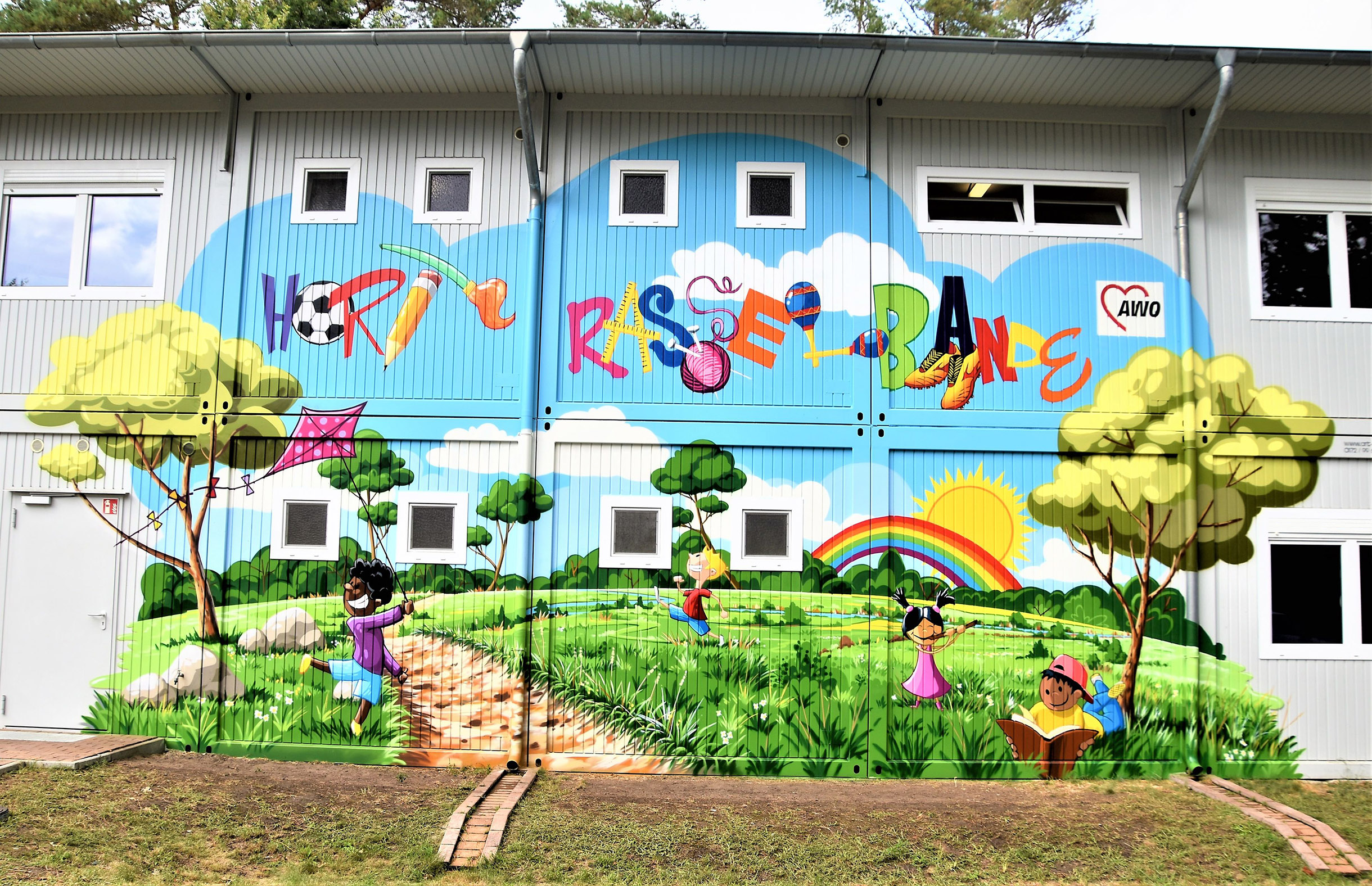 Wandbild an Fassade eines Gebäudes. Bunte Schrift: Hort Rasselbande. Spielende Kinder, Regenbogen, Sonne, Wiese Bäume