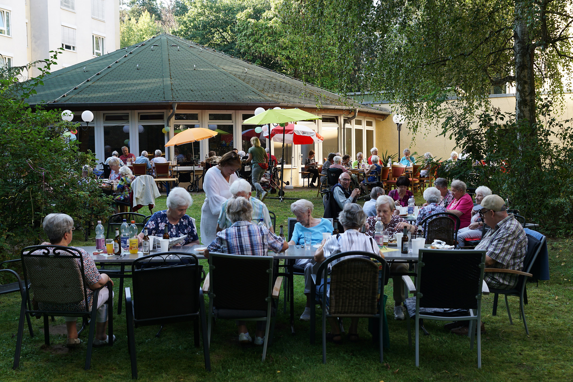 Draußen, Tische sind aneinander gereiht. Ältere Menschen sitzen essen und trinken. Ein großer Baum spendet Schatten, sonst sind kleine farbige Sonnenschirme aufgestellt.