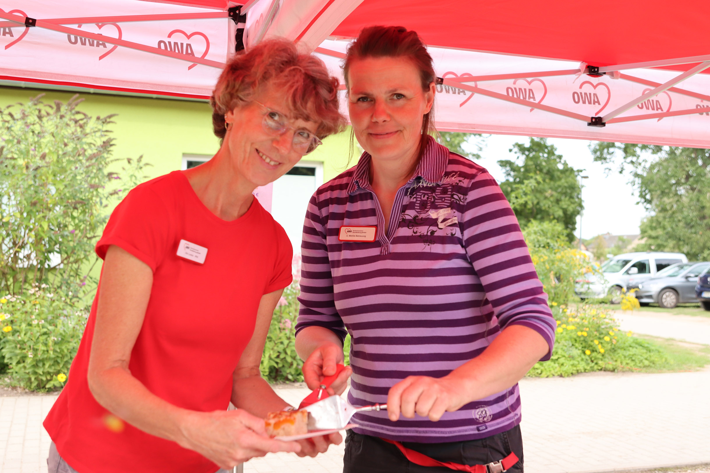 Zwei Frauen unter einem AWO Pavillon. Mit einem Tortenheber tuen sie Kuchen auf. Schönes Wetter. Eine Frau trägt Brille und ein rotes T-Shirt. Die andere hat braune zurück gekämmte Haare und ein Lila gestreiftes Poloshirt an.