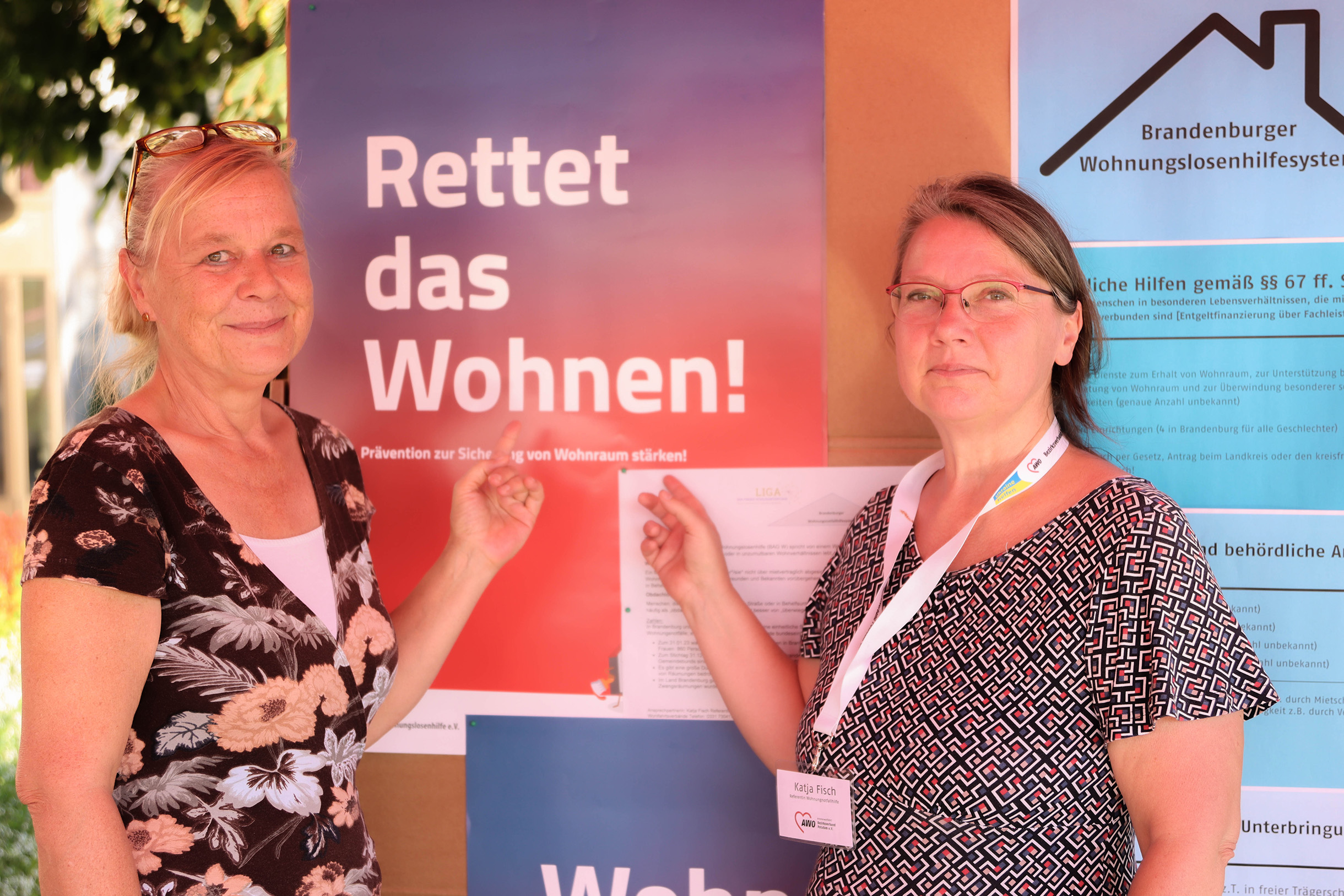Zwei Frauen stehen vor einer Info-Tafel. An der Tafel sind Informationen zum Thema wohnen vom Brandenburger Wohnungslosenhilfesystem. Beide zeigen auf ein Plakat Text: Rettet das Wohnen!