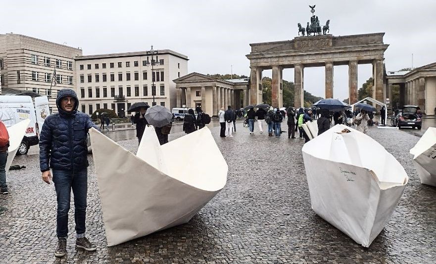 Vorplatz des Brandenburger Tor in Berlin. Regenwetter. Menschen mit Regenschirmen und Kapuzenjacken. 2 XXL-Origami-Papierboote stehen auf dem Platz. Ein Mann steht neben einem der Boote, er trägt eine dunkelblaue Regenjacke die Kapuze hat er aufgesetzt.