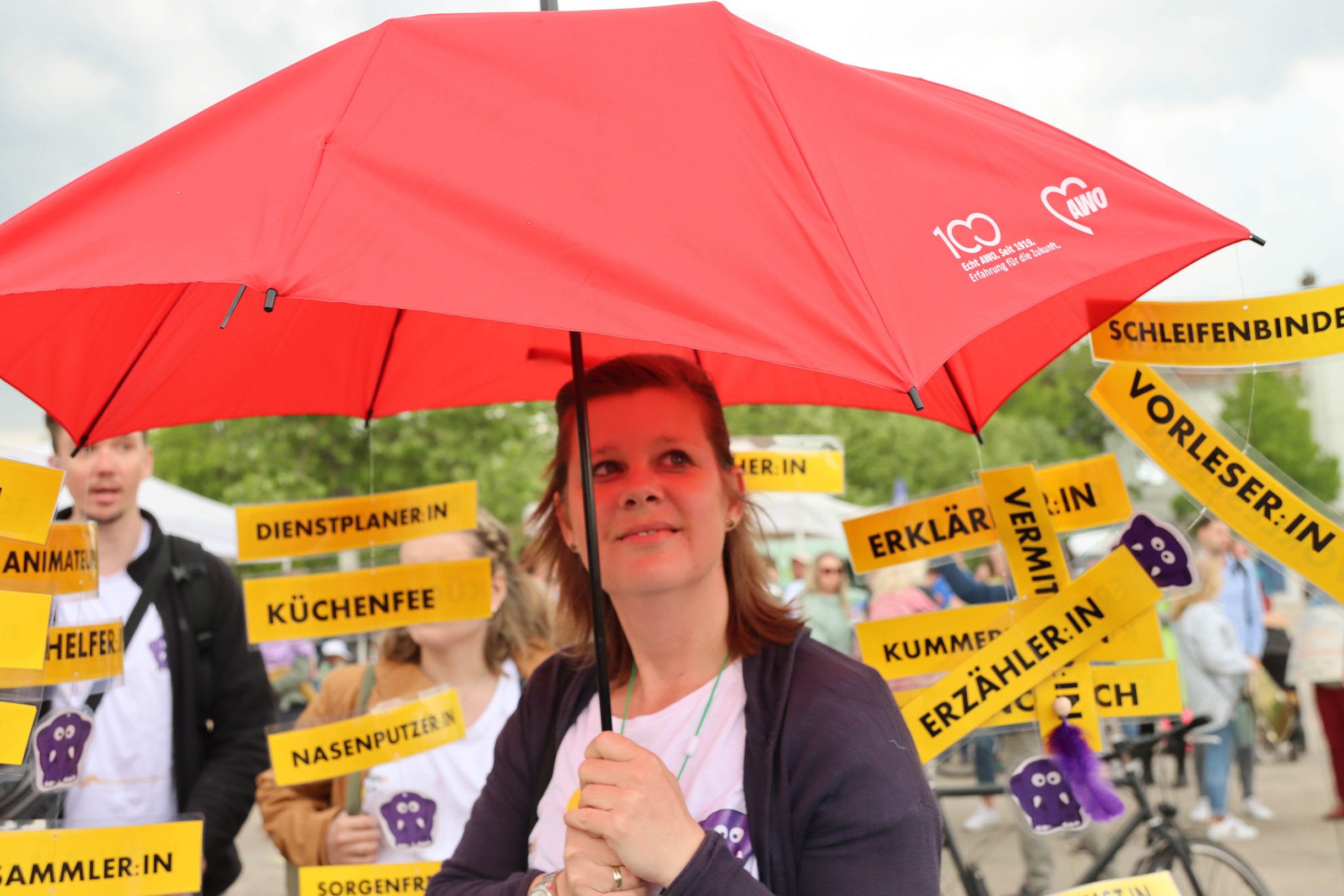 Frau mit großem roten AWO-Regenschirm. Im Hintergrund Menschen mit gelben Schildern. Text auf verschiedenen Schilder: Vorleser, Schleifenbinder, Erzähler Dienstplaner, Küchenfee
