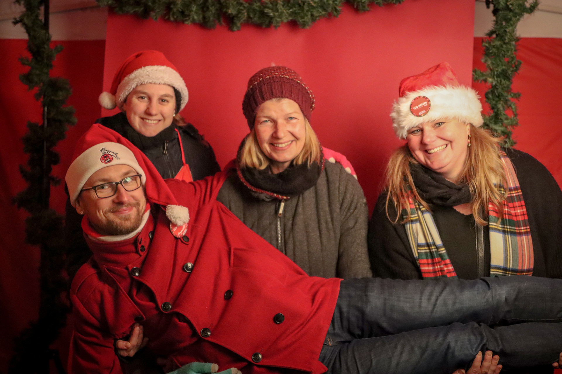 Drei Frauen lächeln, ein Mann liegt über deren queer auf ihren Beinen. Sie haben rote Weihnachmützen auf. Der Mann trägt eine Brille. Sie posieren vor einem roten Hintergrund.