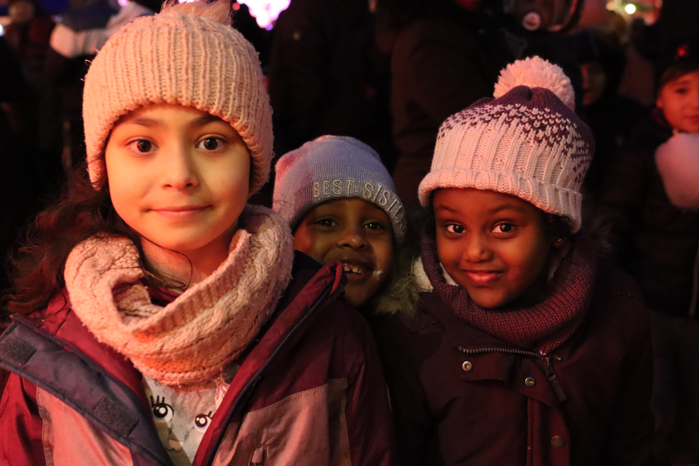 Drei Mädchen. Mit Winterkleidung, Mützen, Schal, Winterjacke. Im Alter von ca. 5 - 7 Jahren. Zwei der Mädchen sind afroafrikanisch (People of Color). Alle drei lächeln fröhlich.