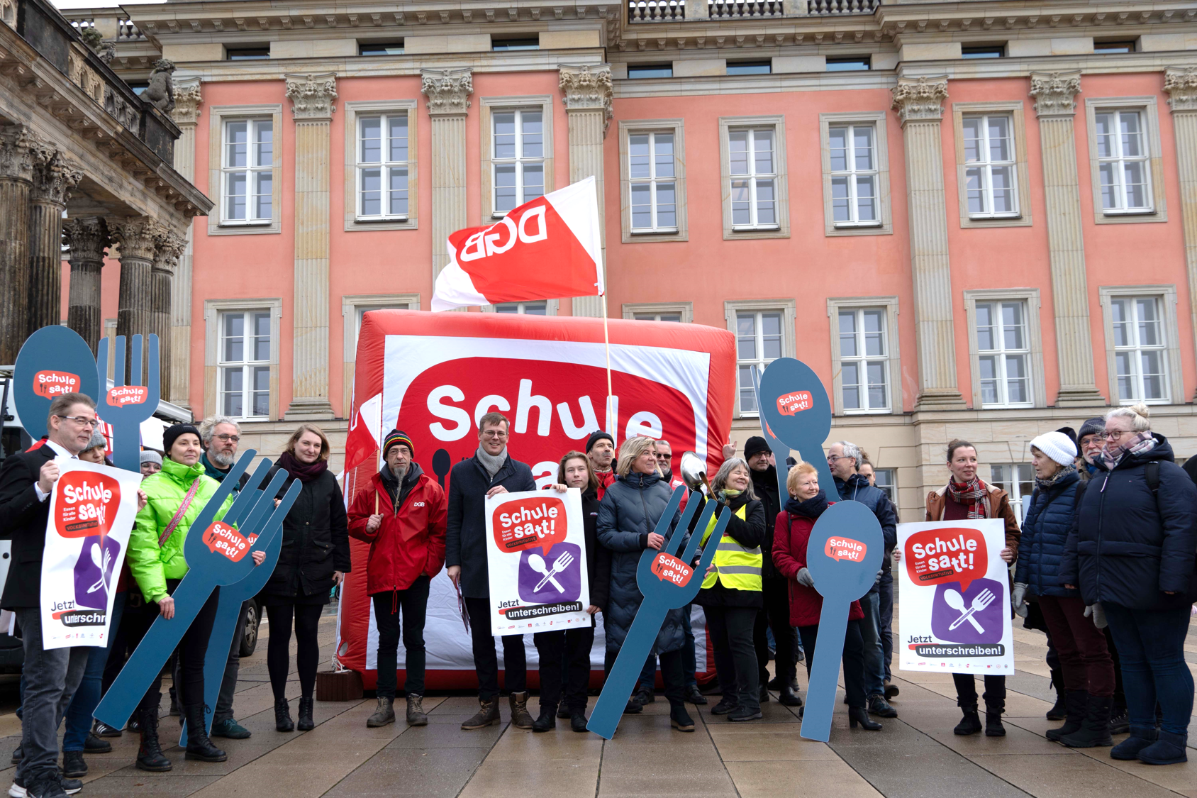Gruppenbild. Vor dem Landtag Brandenburg. Mehrere Personen mit Schildern und großen Gabeln und Löffeln, diese sind beschriftet mit: Schule satt!