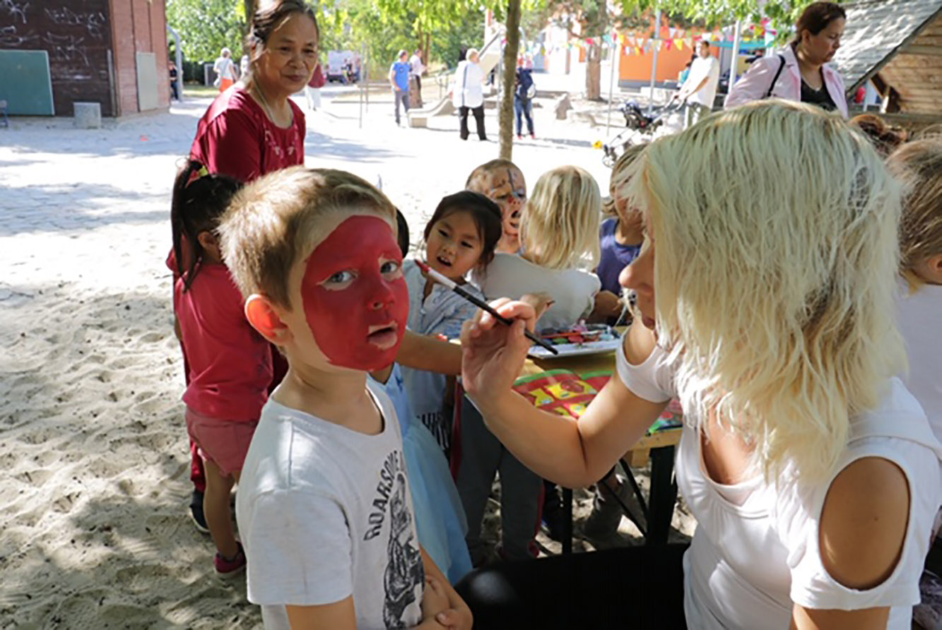 Eine blonde Frau mit schulterlangen blonden Haaren und weißem T-Shirt schminkt einen kleinen Jungen das Gesicht rot. Der Junge trägt ein T-Shirt mit Aufdruck. Im Hintergrund sind noch mehr Kinder.