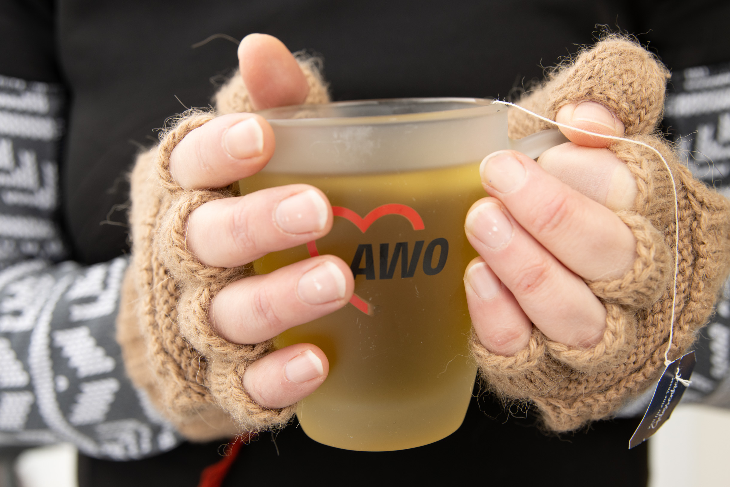 Teetasse mit AWO-Logo. Tasse wird mit zwei Händen in fingerlosen braunen Handschuhen gehalten.