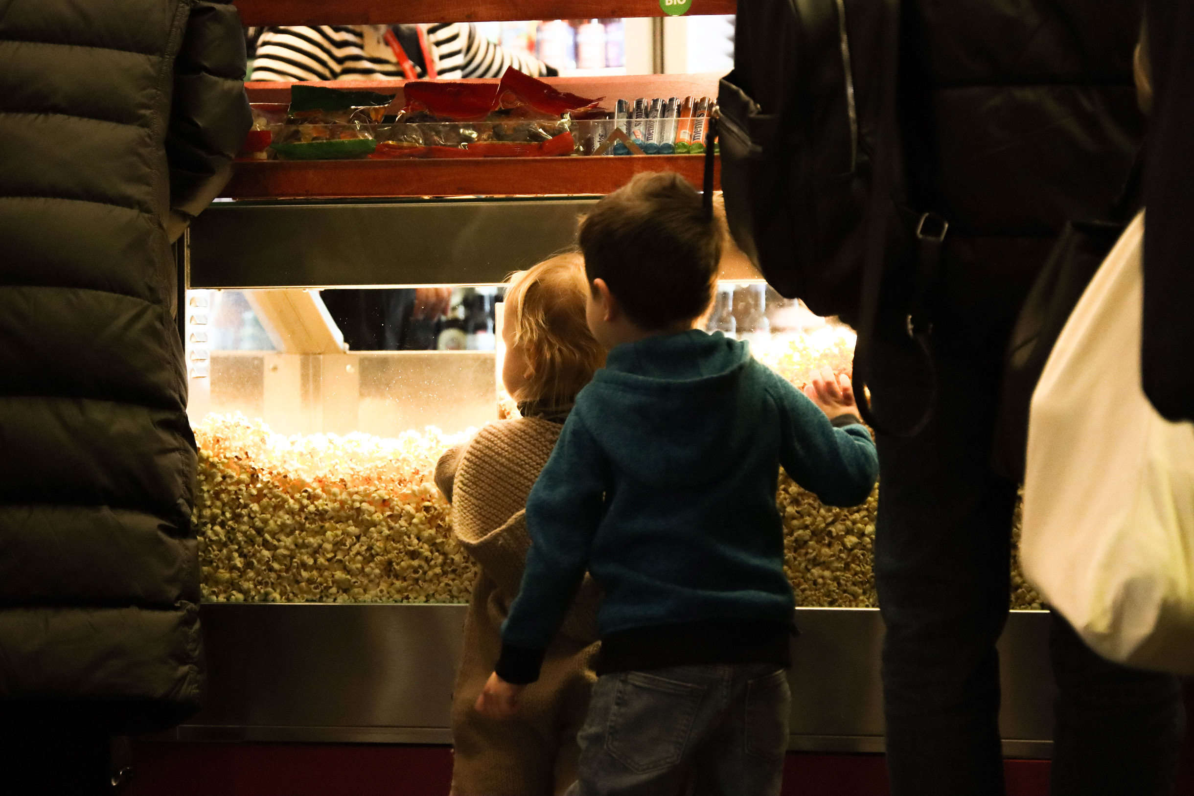 Zwei kleine Kinder stehen vor einem Tresen an dem Popcorn verkauft wird. Von hinten fotografiert. Junge in blauem Pullover/Jacke mit Kapuze. Er hat die Hand an er Scheibe das Popcorn-Verkaufs.