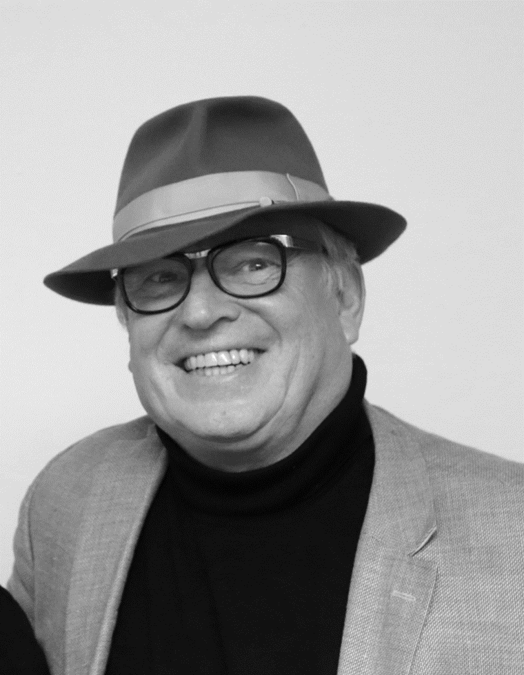 Schwarz-Weiß Portrait: Mann mit Brille und Hut lächelt breit.  Mann ist alt, graue Haare. Volles Gesicht.