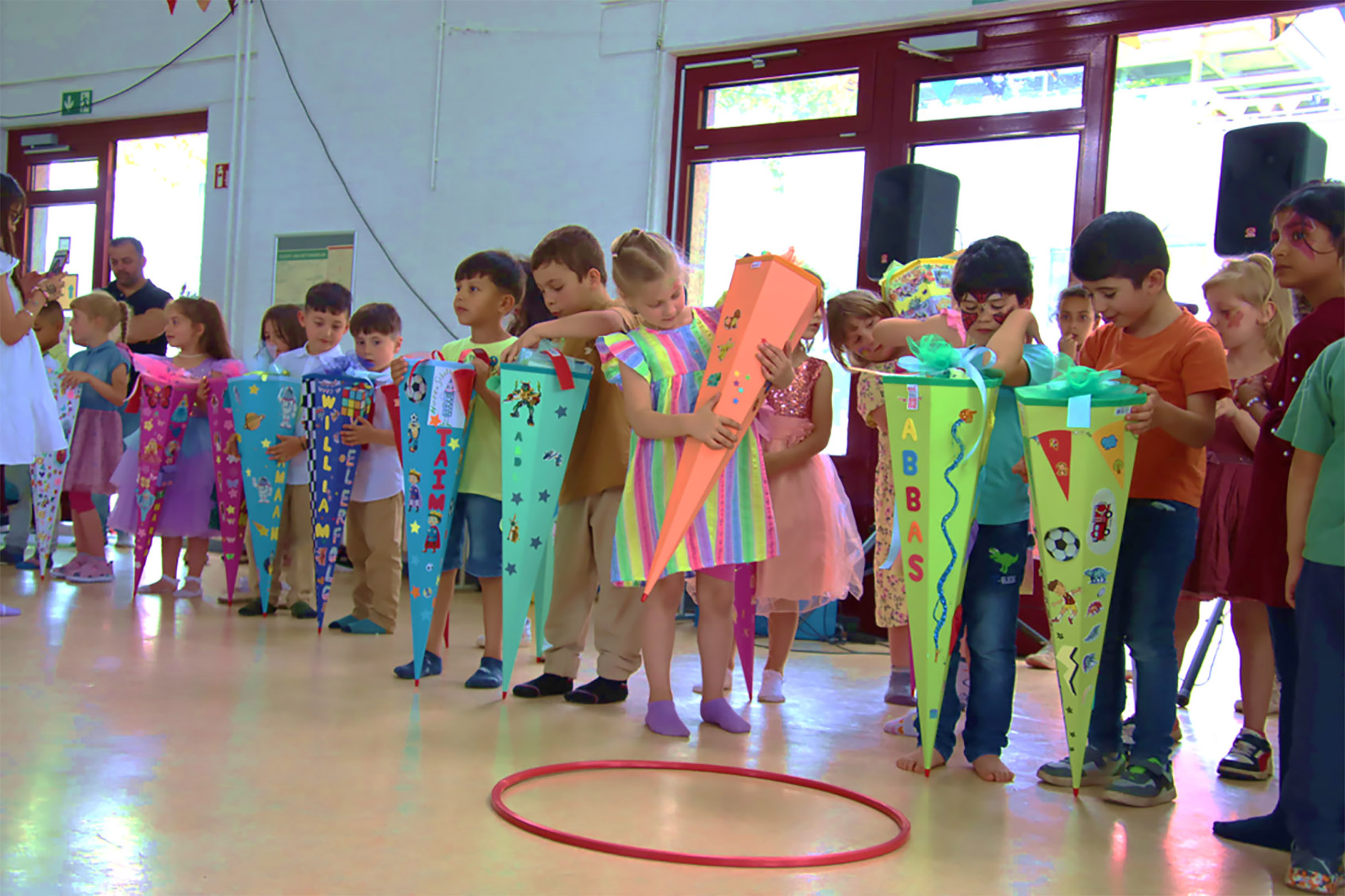 Ca. 12 Einschulungskinder mit ihren großen Zuckertüten in einem großen Raum. Die Zuckertüten sind farbig und bund beklebt. Die Kinder freuen sich.