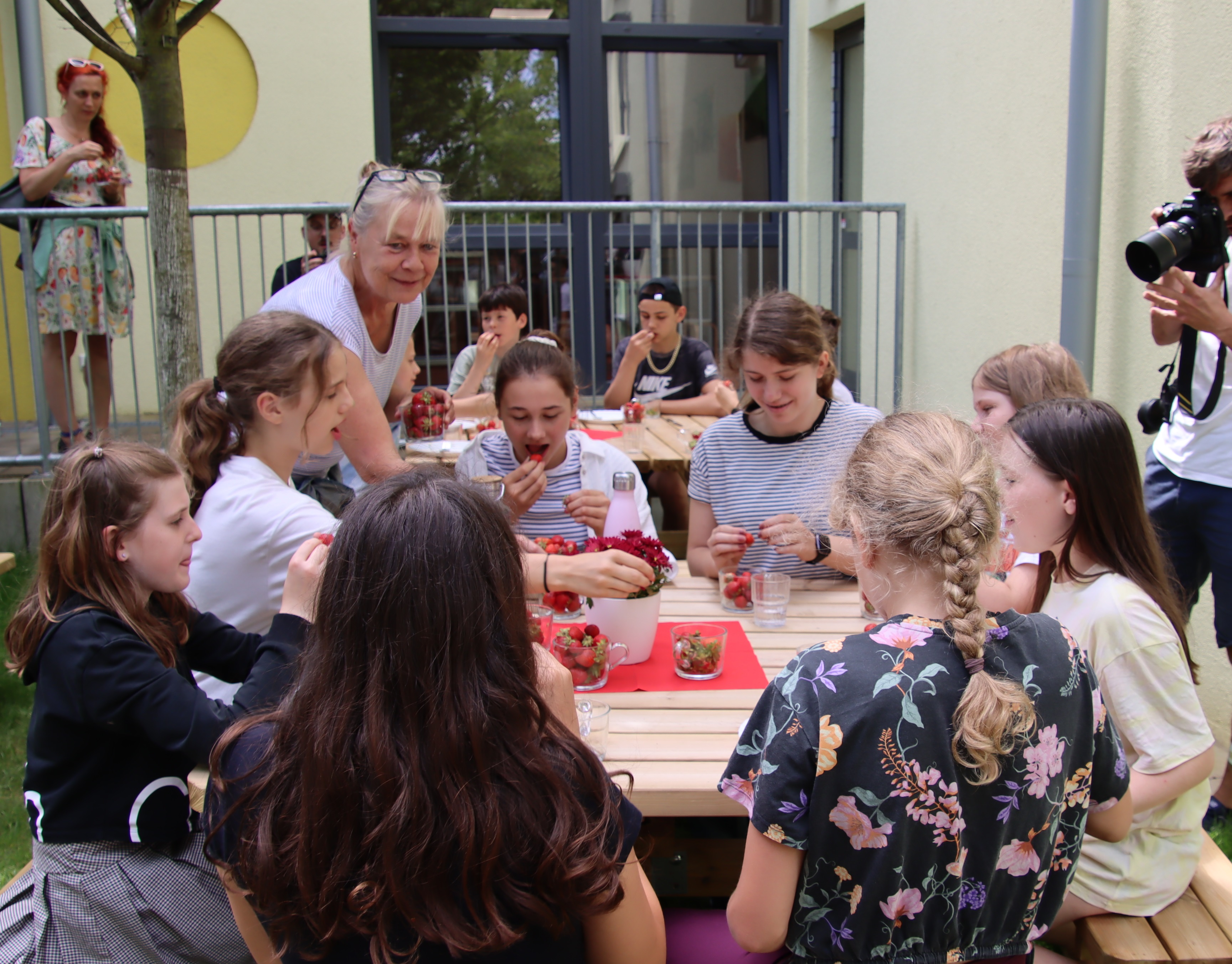 Draußen. Kinder sitzen an Holztischen.Sie essen Kirchen. Ein Fotograf fotografiert die Szene, er steht rechts neben einem Tisch. Eine frau mit blonden Haaren, Ponny, Brille auf den Kopf geschoben steht am Tisch 8 Mädchen drum herum sitzen.