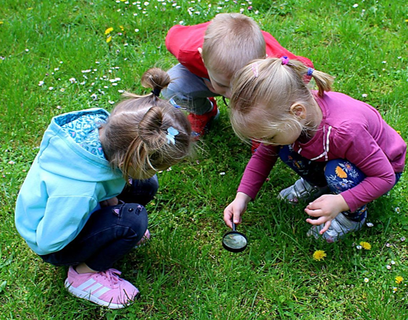 Drei kleine Kinder, 2 Mädchen und ein Junge hocken auf einer Wiese und schauen durch eine Lupe, die das eine Mädchen über das Gras hält