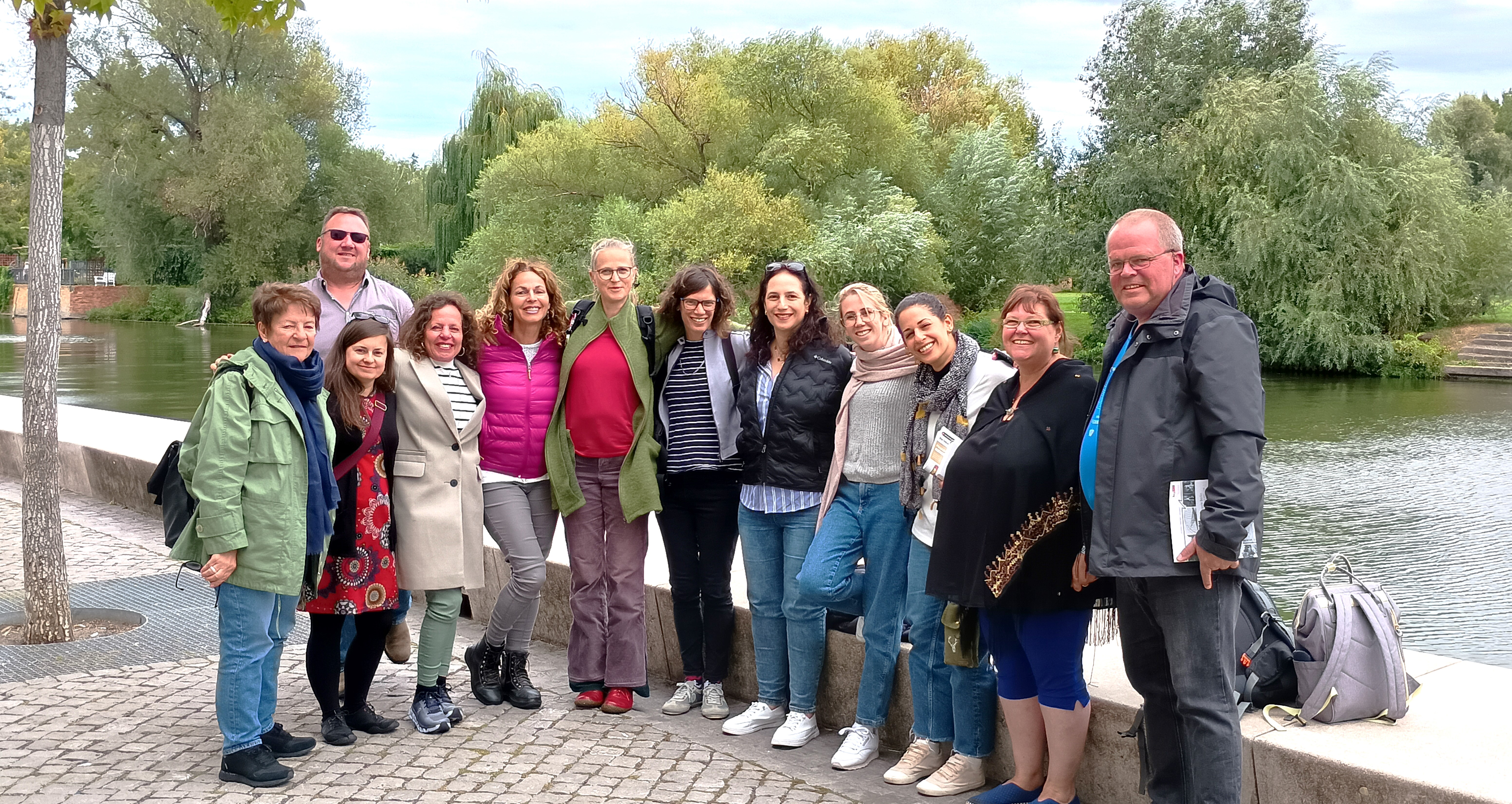 Gruppenbild. 10 Frauen, 2 Männer stehen an einem Fluss. Bäume und viel grün im Hintergrund auf der anderen Flussseite.