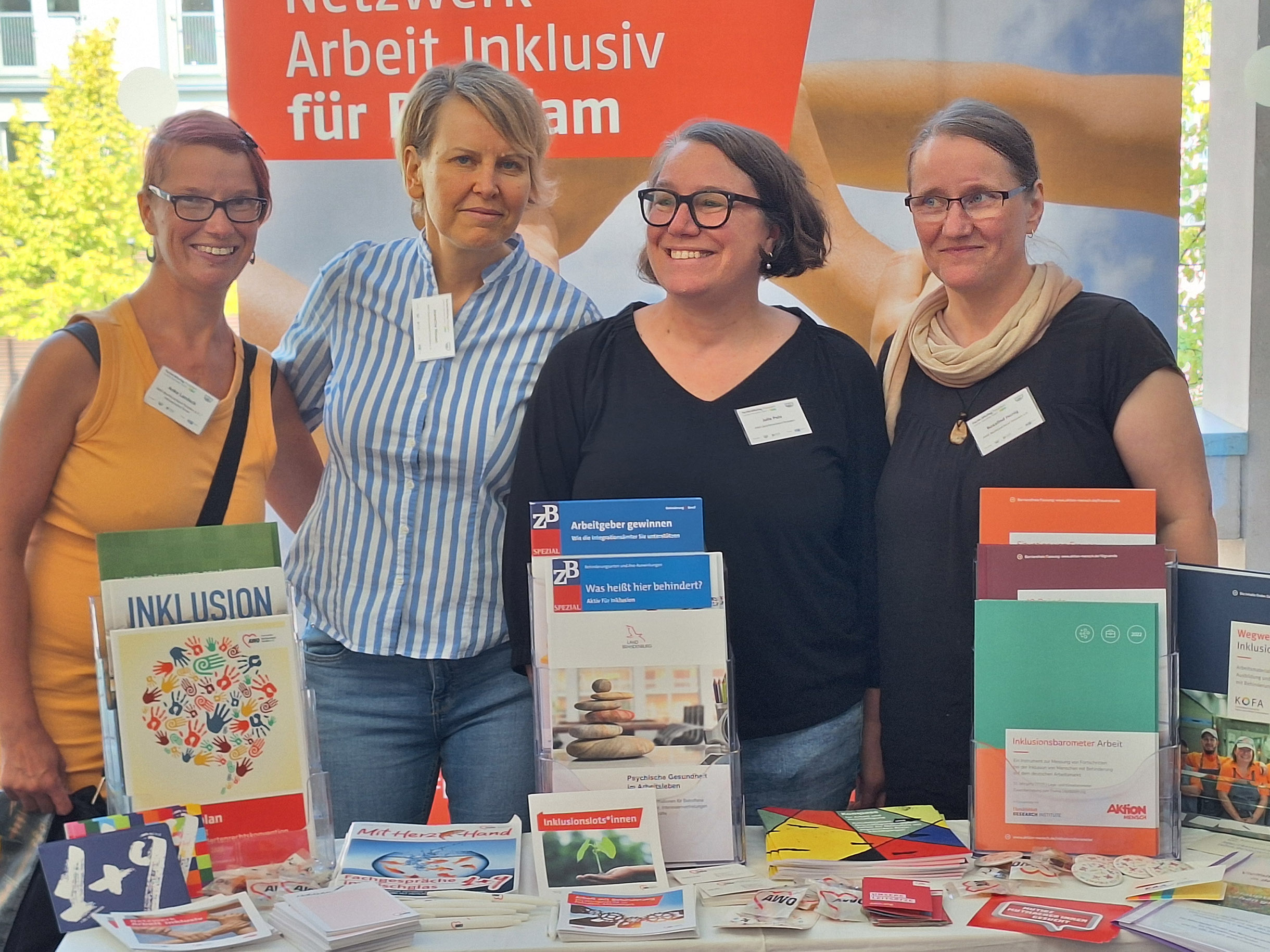 4 Frauen stehen nebeneinander am Stand des Netzwerk Arbeit inklusiv und lächeln in die Kamera. Vor ihnen auf dem Tisch Informationsmaterial zum Thema Arbeit mit Behinderung.