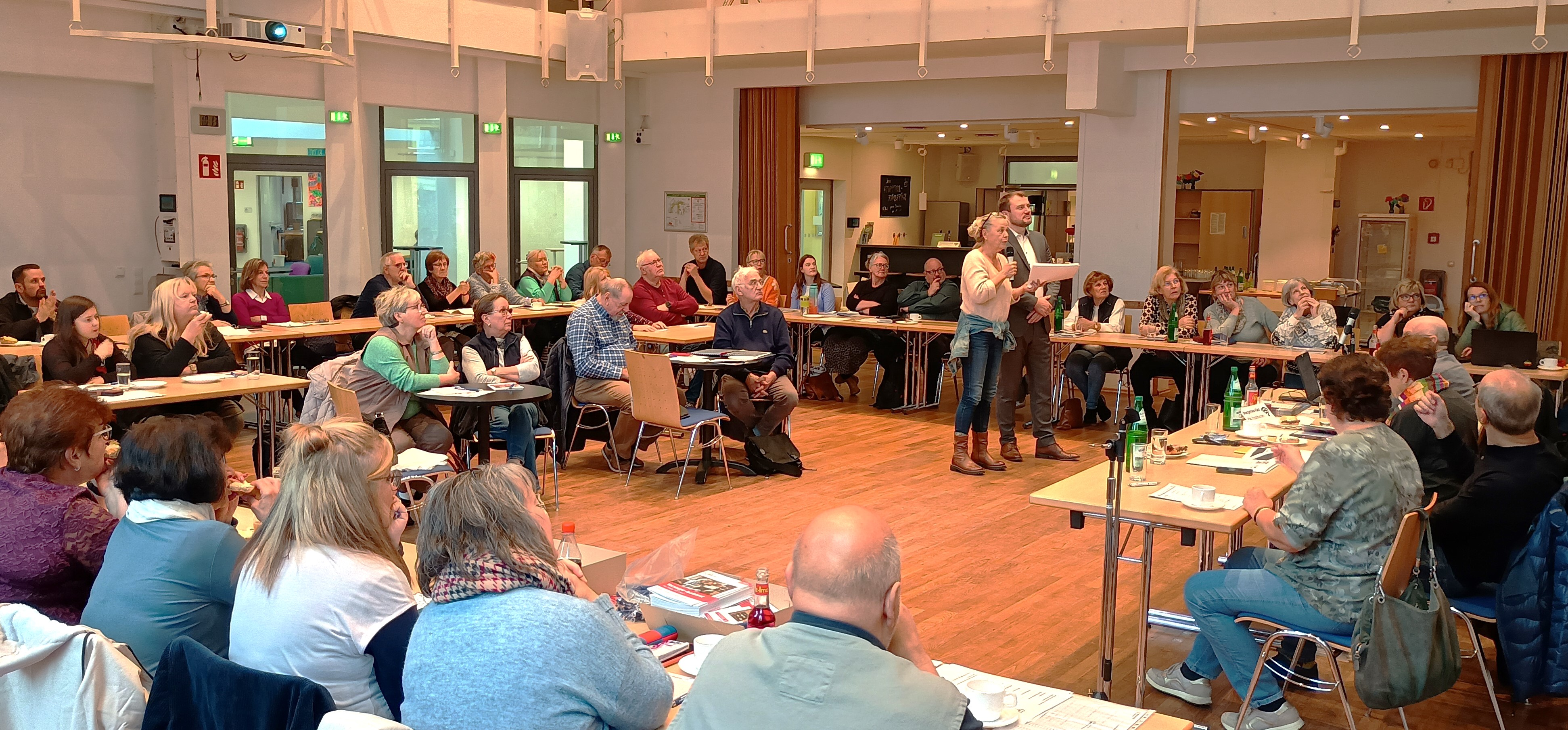 Sehr großer Raum, Tische zu einem großen Viereck gestellt an denen vielen Menschen sitzen. In deren Mitte stehen eine Frau und mit Mikro und ein Mann.