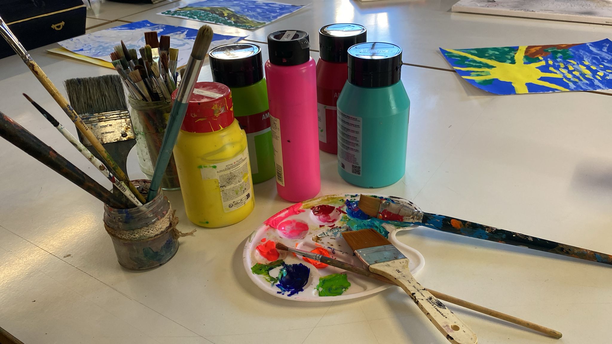verschiedene Tuben Farbe und ein Glas mit Pinsenl stehen auf einem Tisch. Daneben ist eine Mischpalette.