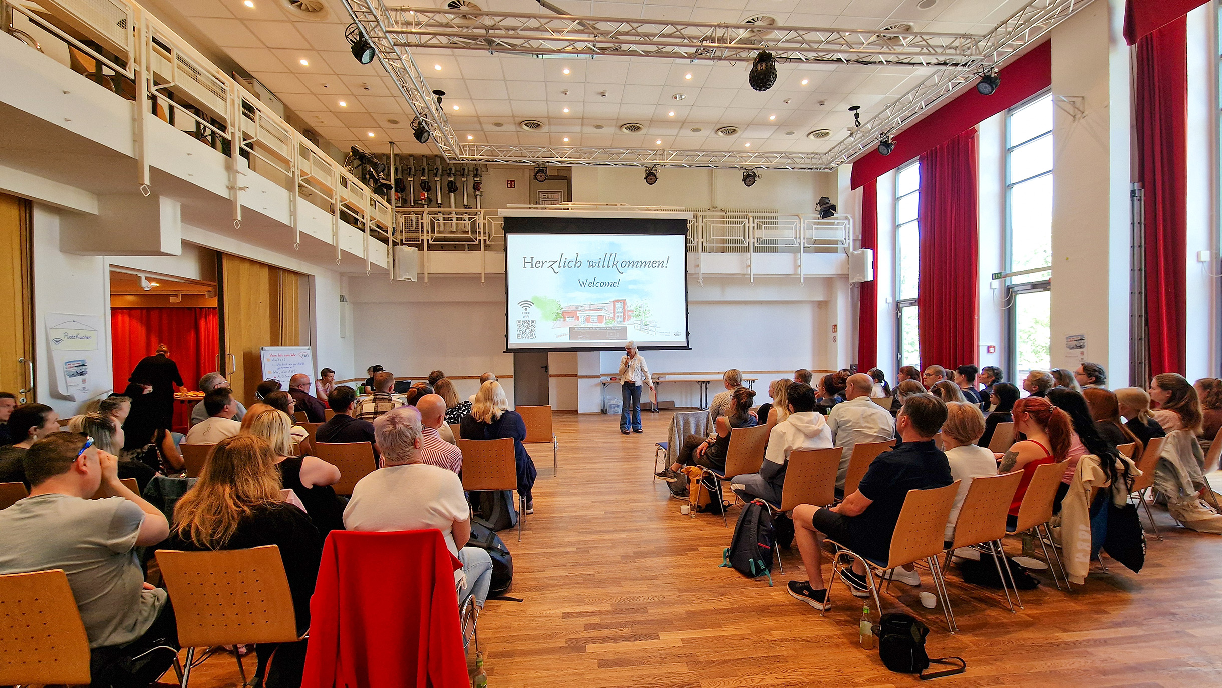 Großer heller Veranstaltungsraum mit vielen Personen, die auf Stühlen sitzen und einer Präsentation auf einer Leinwand folgen. Vorne steht eine Person am Rednerpult.