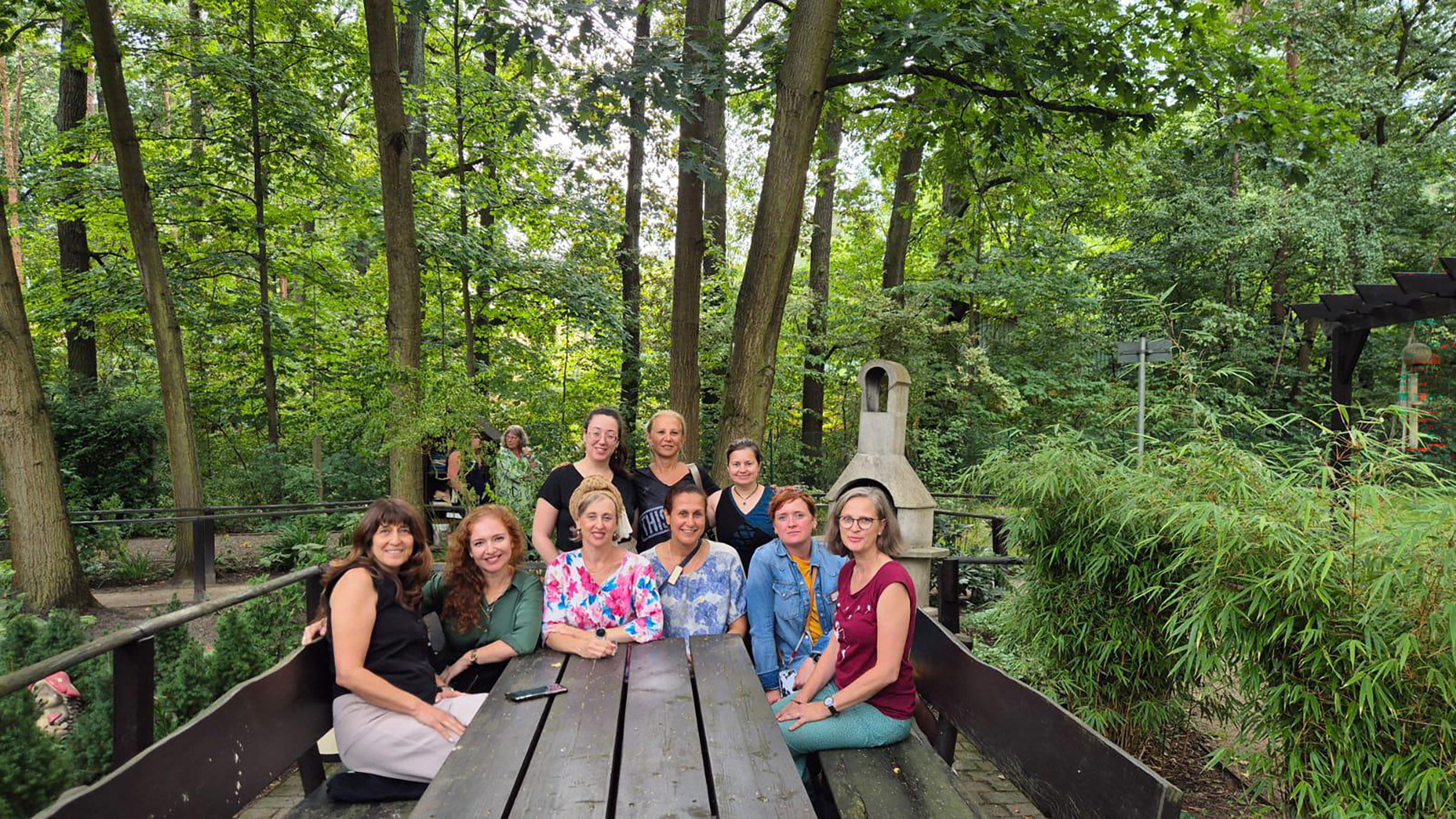 Gruppenfoto: 9 Personen sitzen im Halbrund an einem Holztisch, der in einem bewaldeten Gelände steht. Alle sehen in die Kamera.