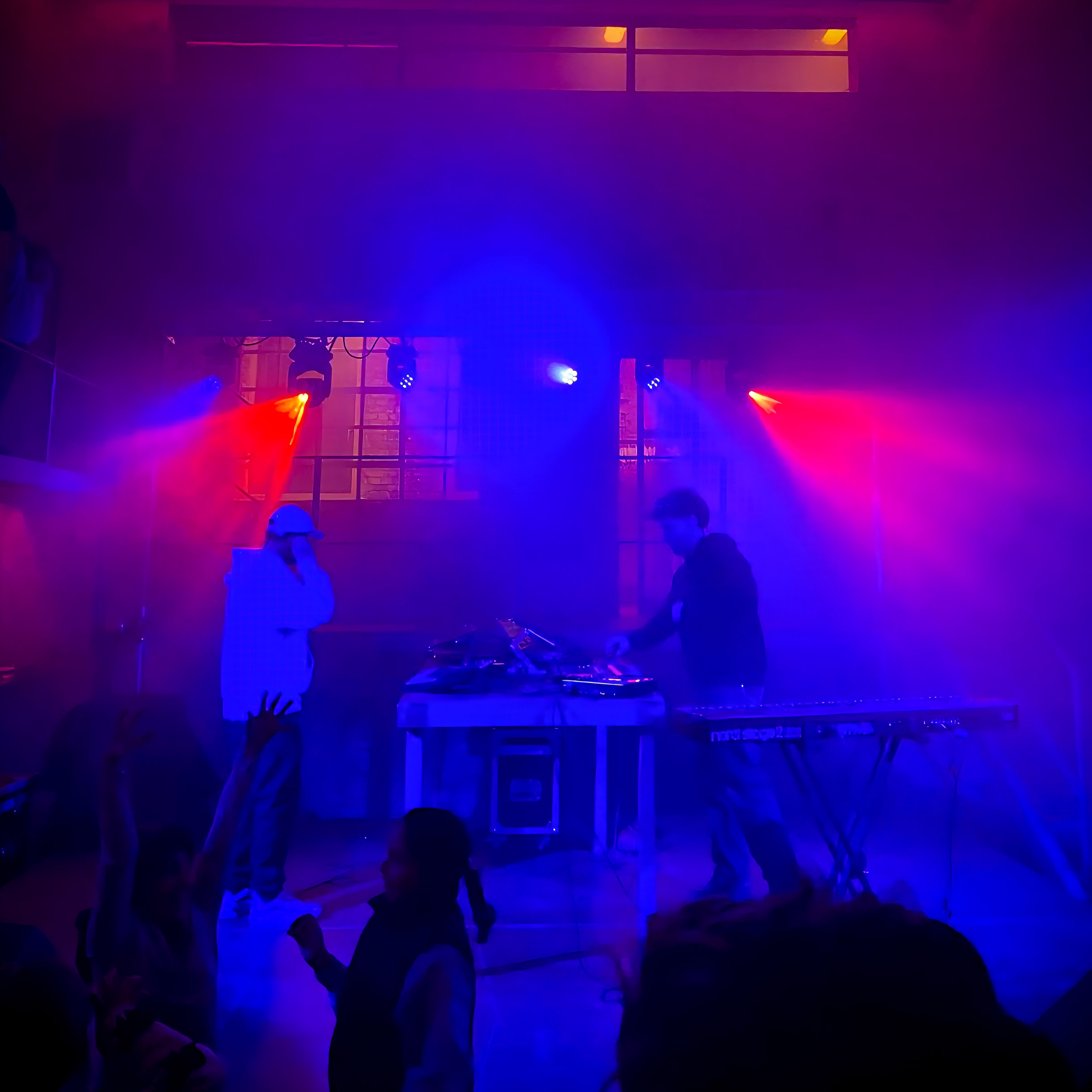 DJs am Pult, es ist dunkel, rot blaues Scheinwerferlicht sorgt für Party-Feeling. Tanzende Menschen im Vordergrund.