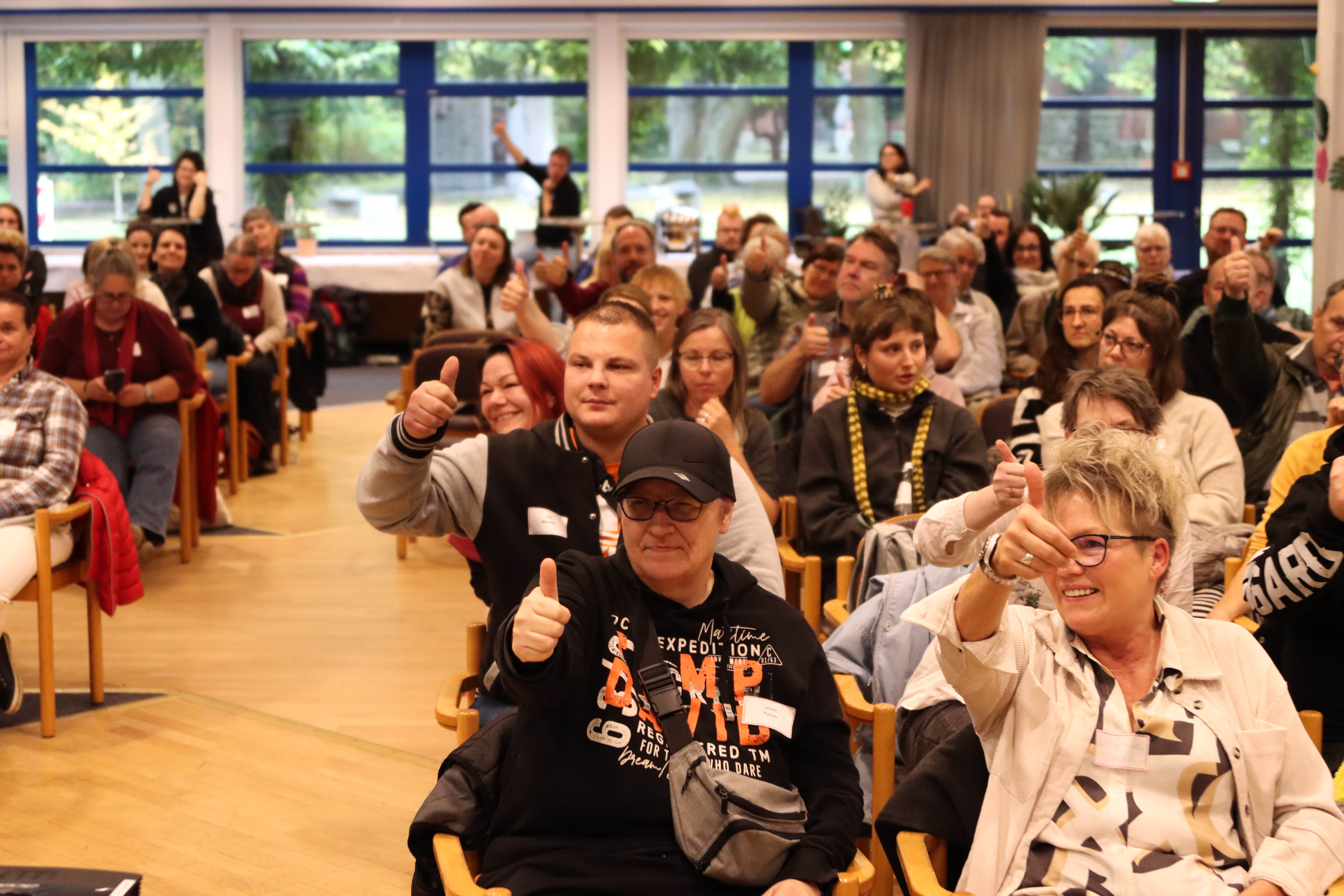 Großer Raum, Viele Teilnehmende sitzen auf Stühlen, lächeln und strecken den Daumen nach oben.
