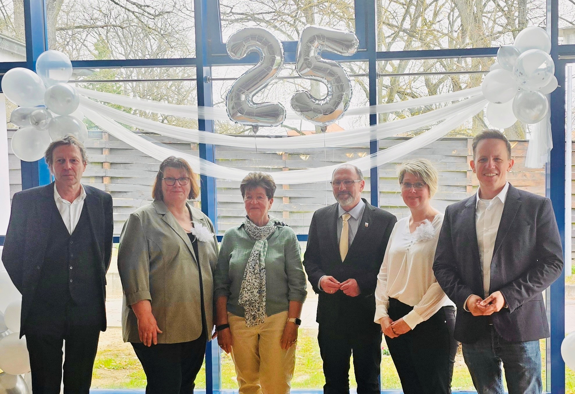 Gruppenbild mit Bürgermeister. 3 Frauen, 3 Männer vor einer Glaswand. Blick nach draußen. Über Ihnen hängt eine silberne Luftballon-25 und weiße Luftballons links und rechts..