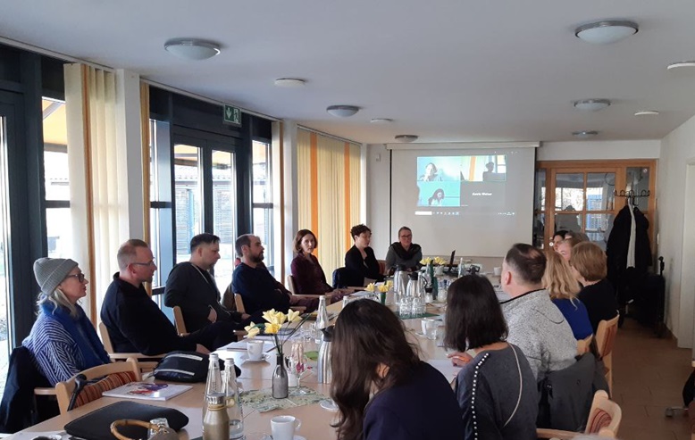 Großer Raum mit einer langen Kaffeetafel, ca. 16 Menschen sitzen am Tisch. Ganz vorn ist eine Leinwand wo Leute über Online Meeting dazu geschalten sind.