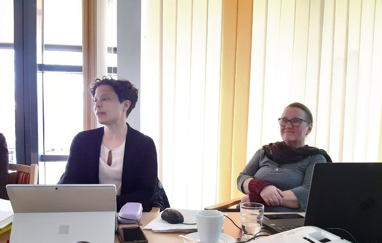 Zwei Frauen sitzen am Tisch. Vor der Frau links ein aufgeklapptes Tablet, vor der anderen Frau ein Laptop.