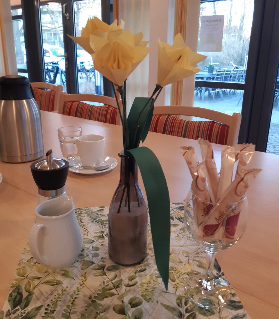 Kaffeetisch, dekoriert mit gebastelten gelben Tulpen aus Papier in einer Vase mit Sand, rechts daneben ein Weinglas mit Amicelli Stangen Schokoröllchen gefüllt.