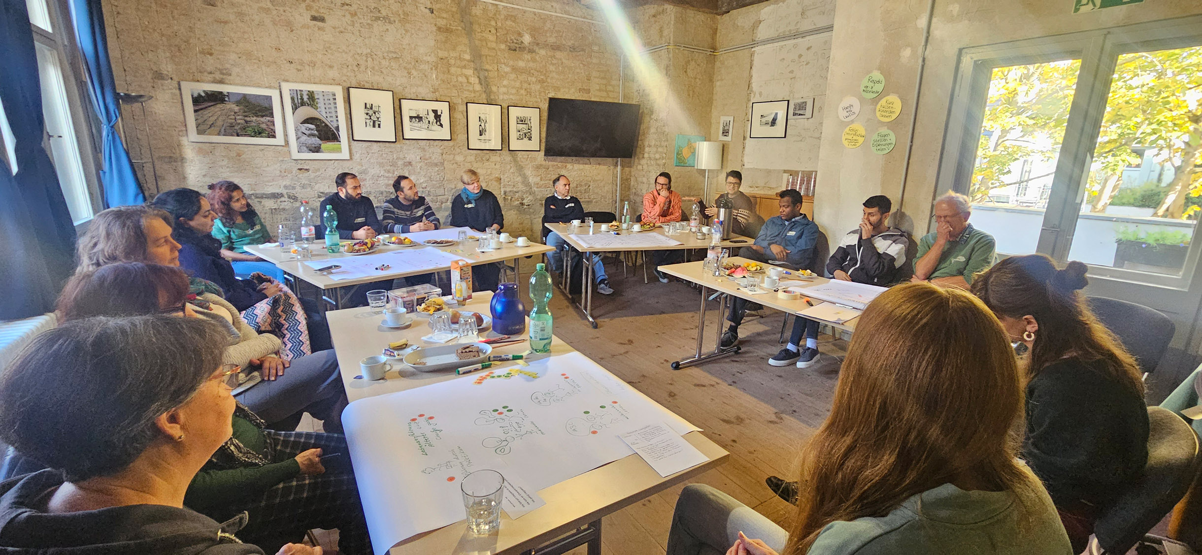 Großer Raum, 16 Teilnehmende sitzen an Tischen in einem Kreis.