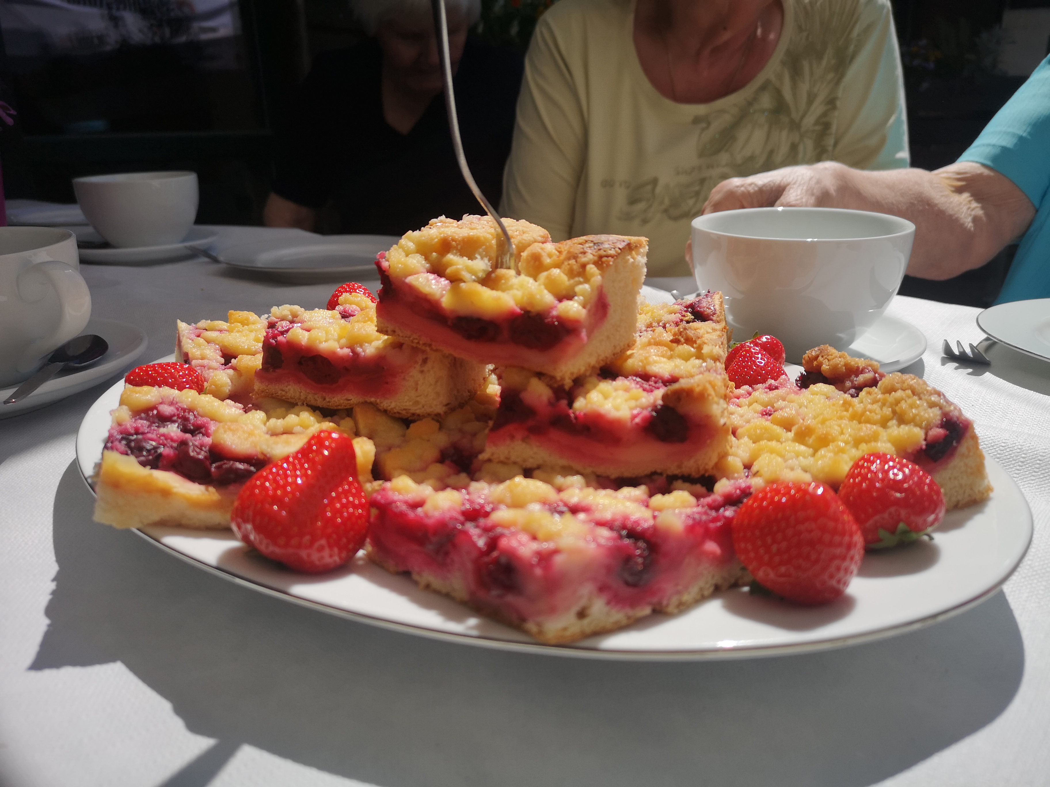 Mehrere Stücke Blechkuchen auf einem weißen Teller mit Erdbeeren. Ein Tisch an dem Menschen sitzen mit Kaffee Geschirr.