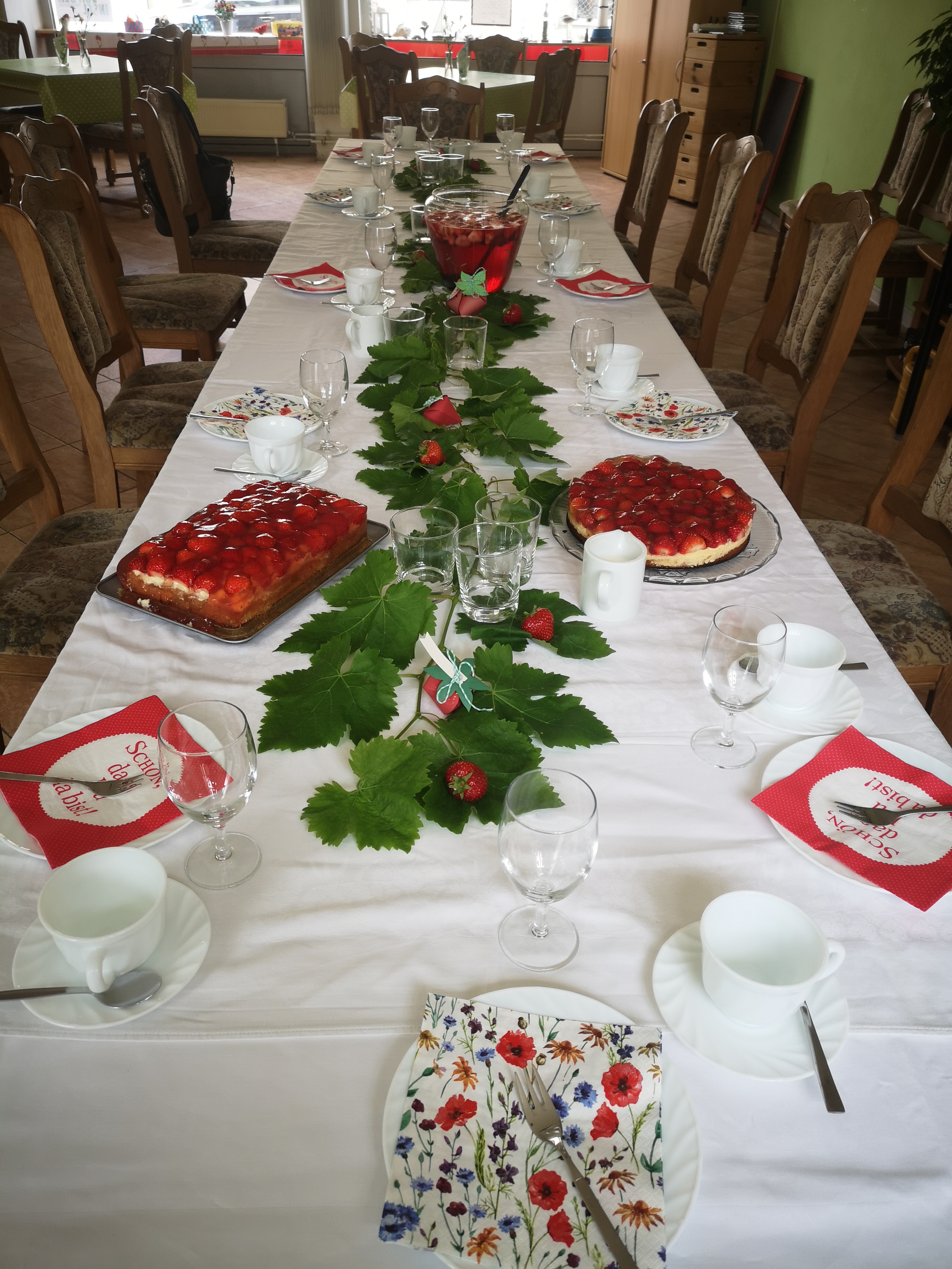 Gedeckte Kaffetafel, Erdbeerkuchen auf dem Tisch. Tisch ist mit Erdbeeren und Erbeerpflanfen-Dekoration geschmückt. Erdbeerbowle steht  in einer Großen Glas-Karaffe in der Mitte der Tafel.