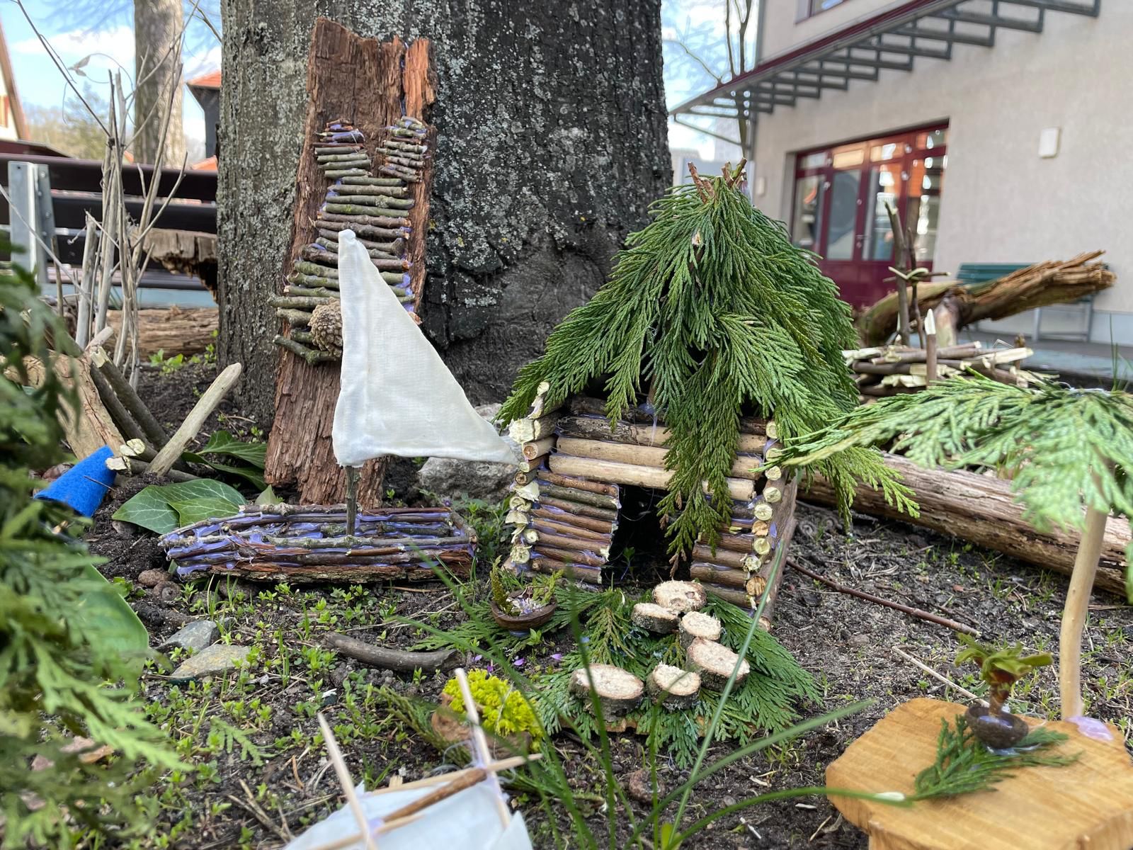 An einem Baum, liegen gebastelte Gegenstände aus Holz und anderen Naturmaterialien. Ein kleines Segelboot, eine Art Hütte mit Tannenzweigen-Dach.