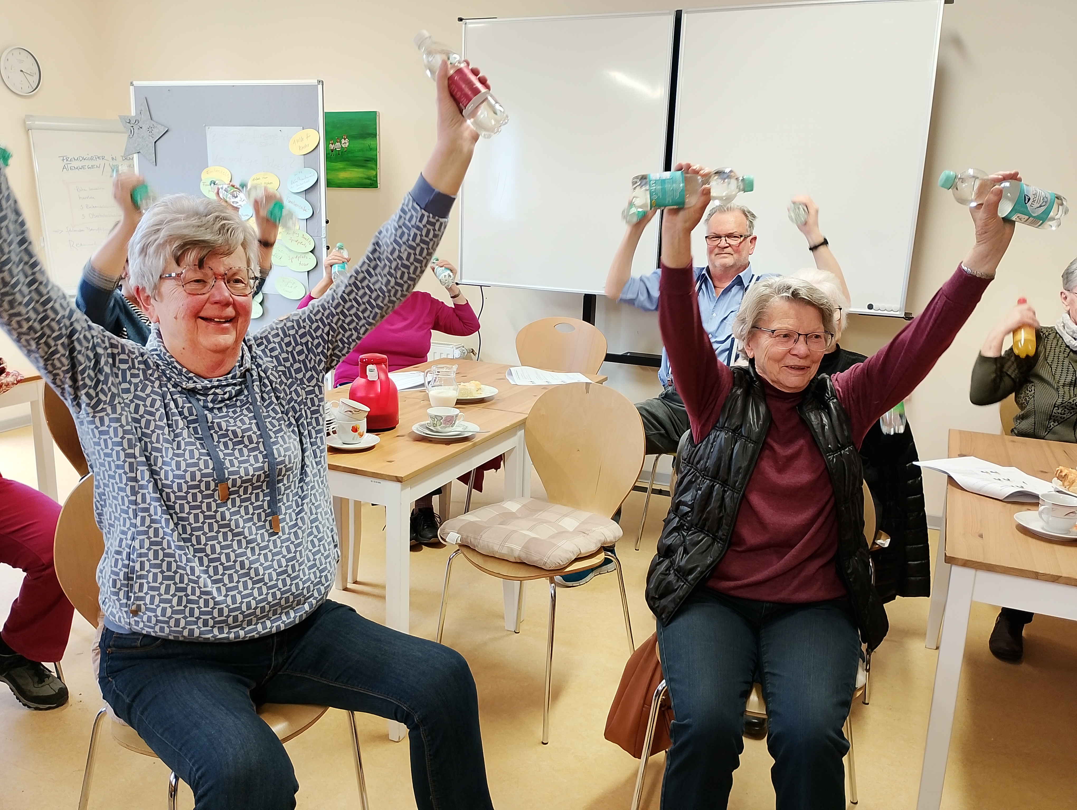 Zwei Seniorinnen sitzen auf Stühlen. Sie haben in jeder Hand eine kleine Flasche Wasser und heben die Arme nach oben. Sie lächeln. Hinter ihnen sitzen vier weitere Senior*inne und machen dieselbe Übung.