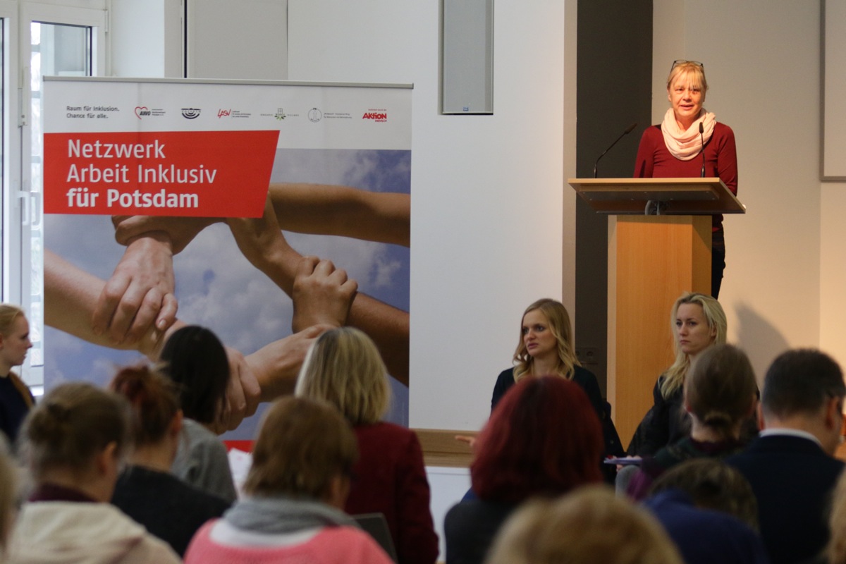 Blonde Frau an einem Rednerpult. Ein großes Plakat vom Netzwerk Arbeit inklusiv für Potsdam. Publikum