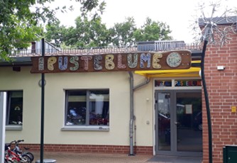 Flachbau mit Fenster und Eingangsbereich, Oben steht in farbigen Buchstaben Pusteblume am Gebäude.
