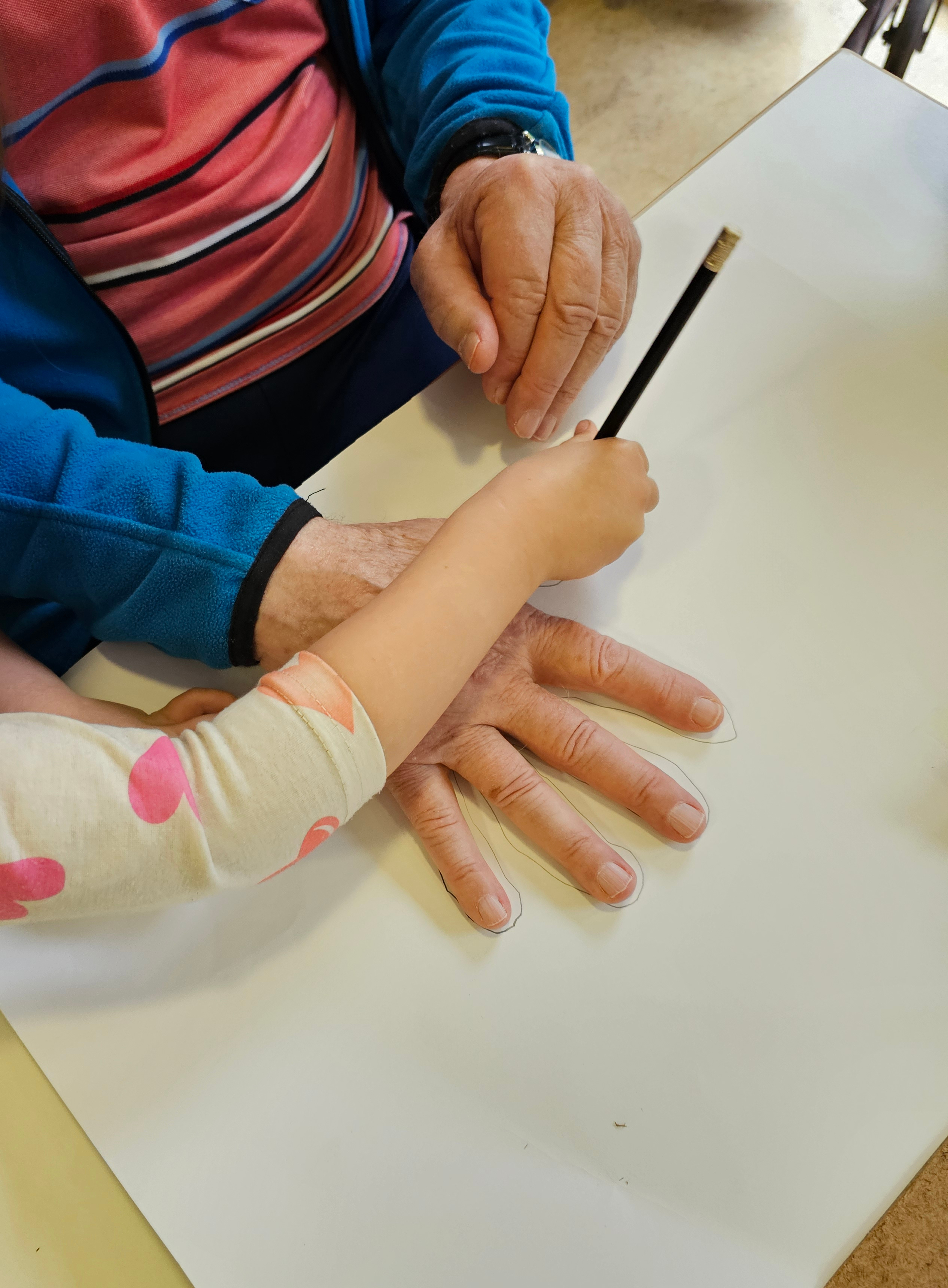 Das Bild zeigt mehrere Hände auf einem Tisch. Die Hände des Kindes halten ein Blatt und umranden die Hand eines älteren Menschen.