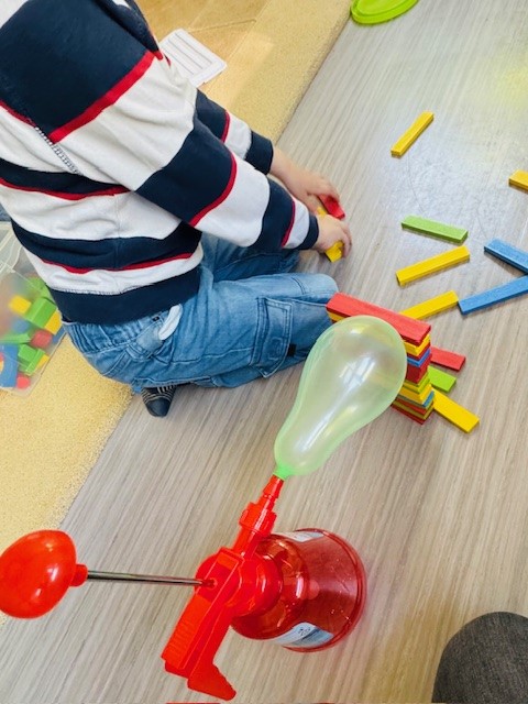 Ein Kind sitzt vor einem Haufen Basteinen. Neben dem Kind steht eine Luftpumpe mit aufgeblasenem Luftballon.