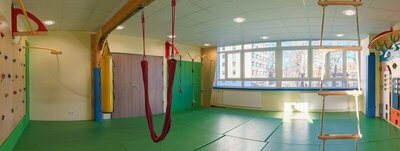 Turnhalle. Grüne Matten am Boden, große Fenster. Klettermöglichkeiten. Hängeleiter.