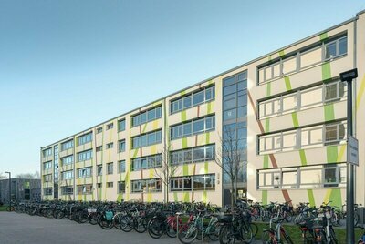 Außenansicht. Sehr großes Gebäude, 4 Stockwerke, langgezogenes Gebäude mit grünlicher Fassade. Viele Fahrräder parken davor.