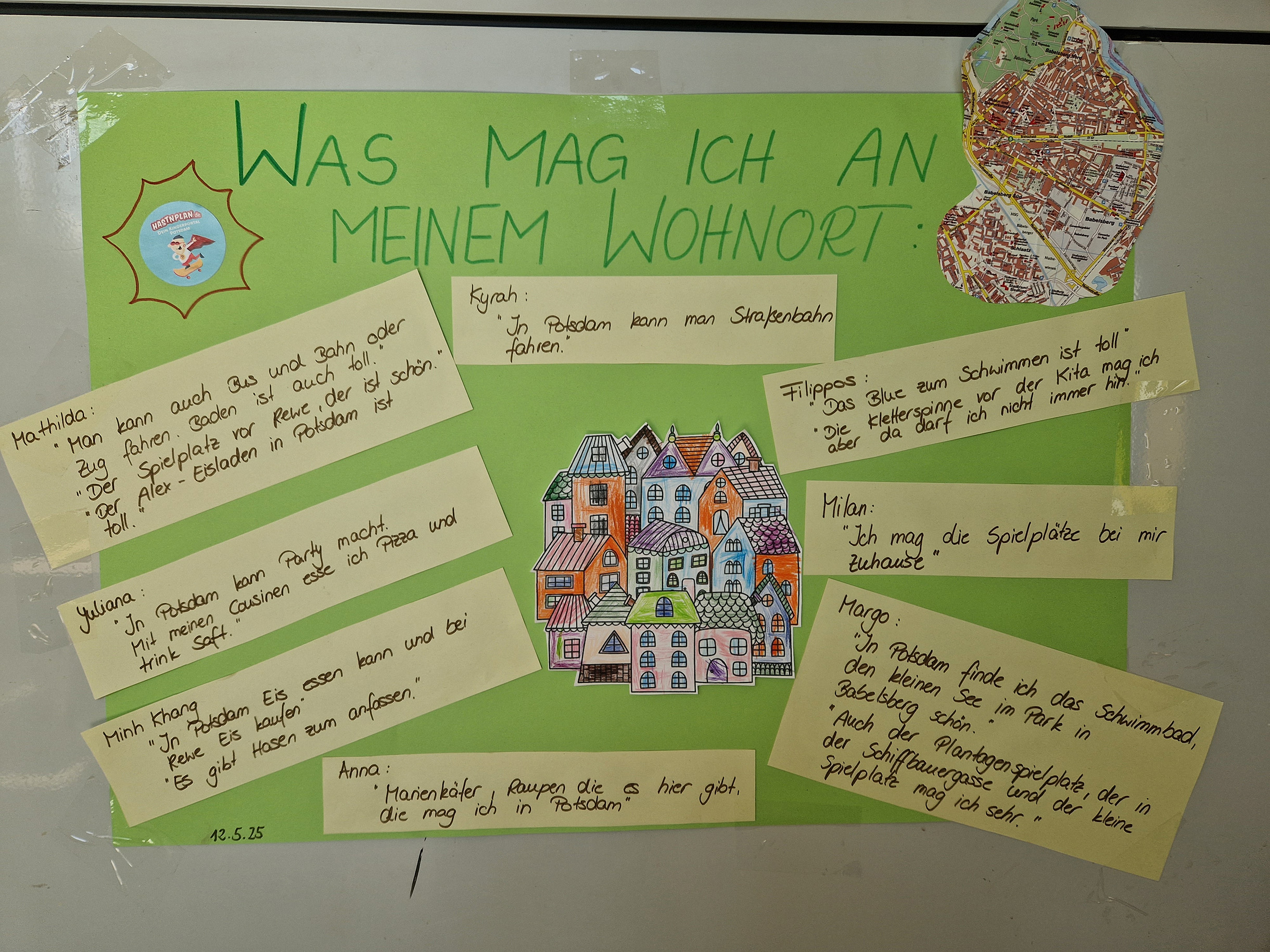 Großes grünes Plakat an einer Wand.  Mit Text: "Was mag ich an meinem Wohnort"  Zitate von Kindern. In der Mitte ein bild mit vielen Häusern.