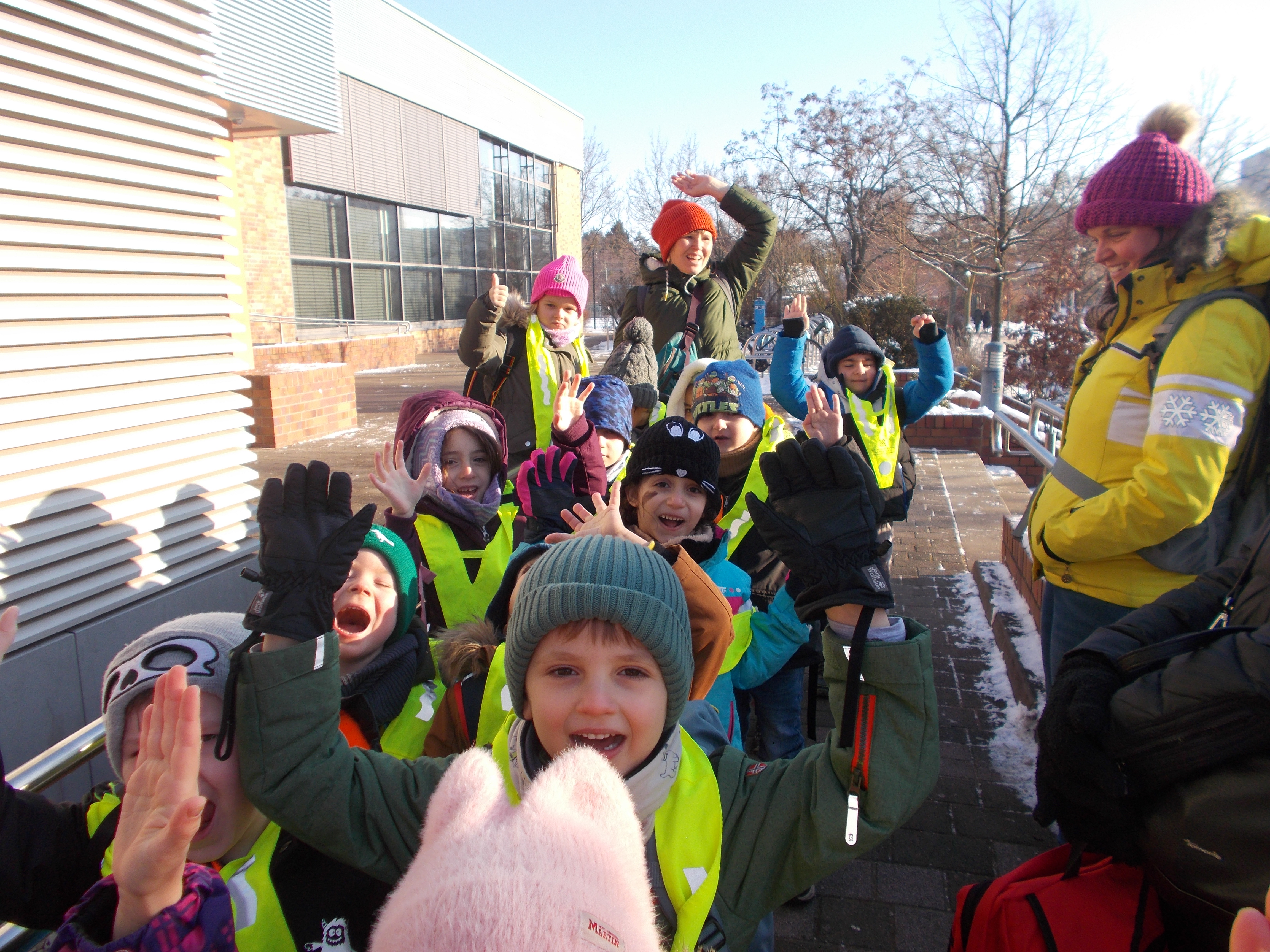 Mehrere Kinder in gelben Warnwesten stehen neben einem Gebäude mit Erzieherin und Eltern. Die Kinder heben vor Freude die Arme und lachen.