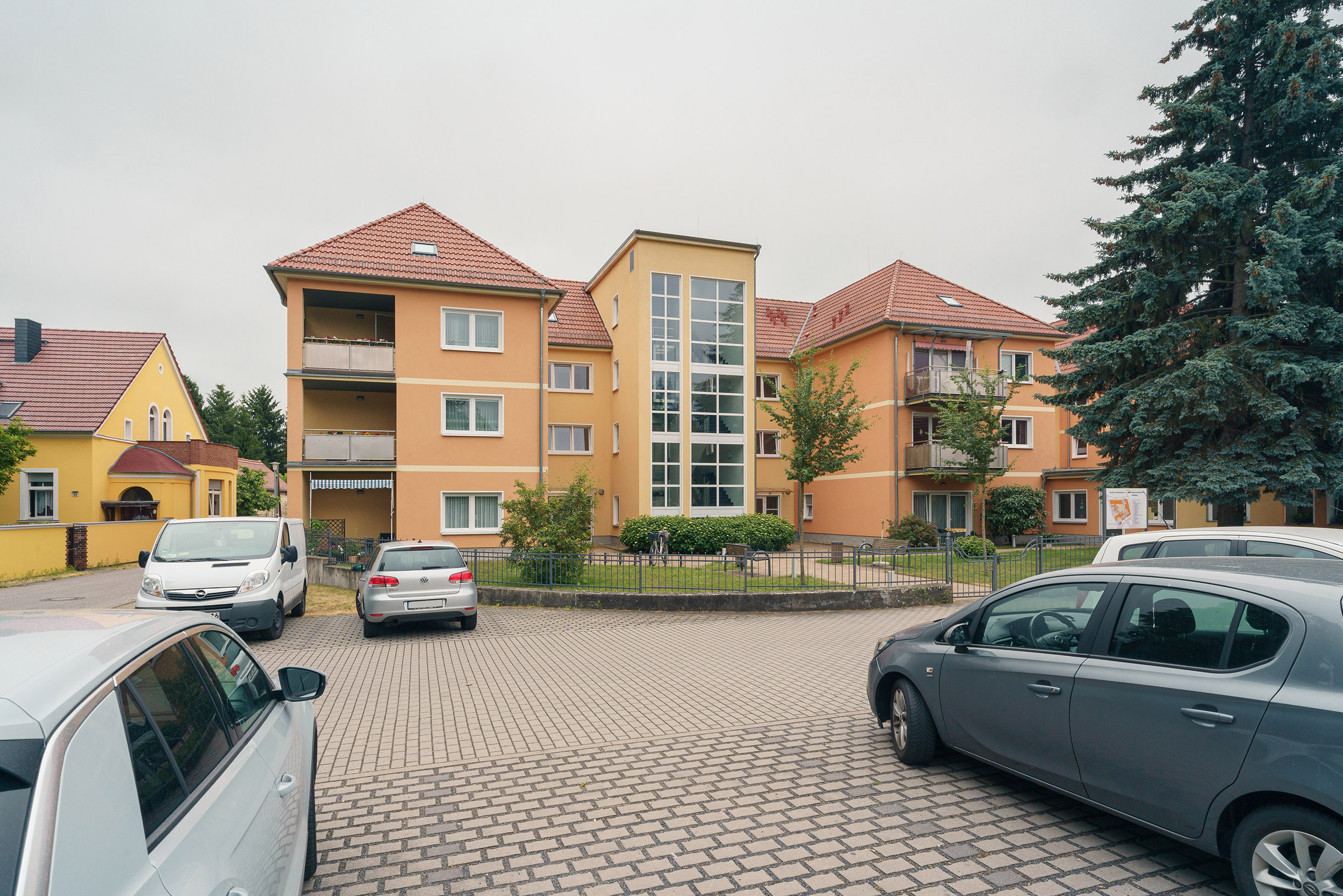 Parkplatz, parkende Autos, ein orangenes Gebäude mit Vorgarten.