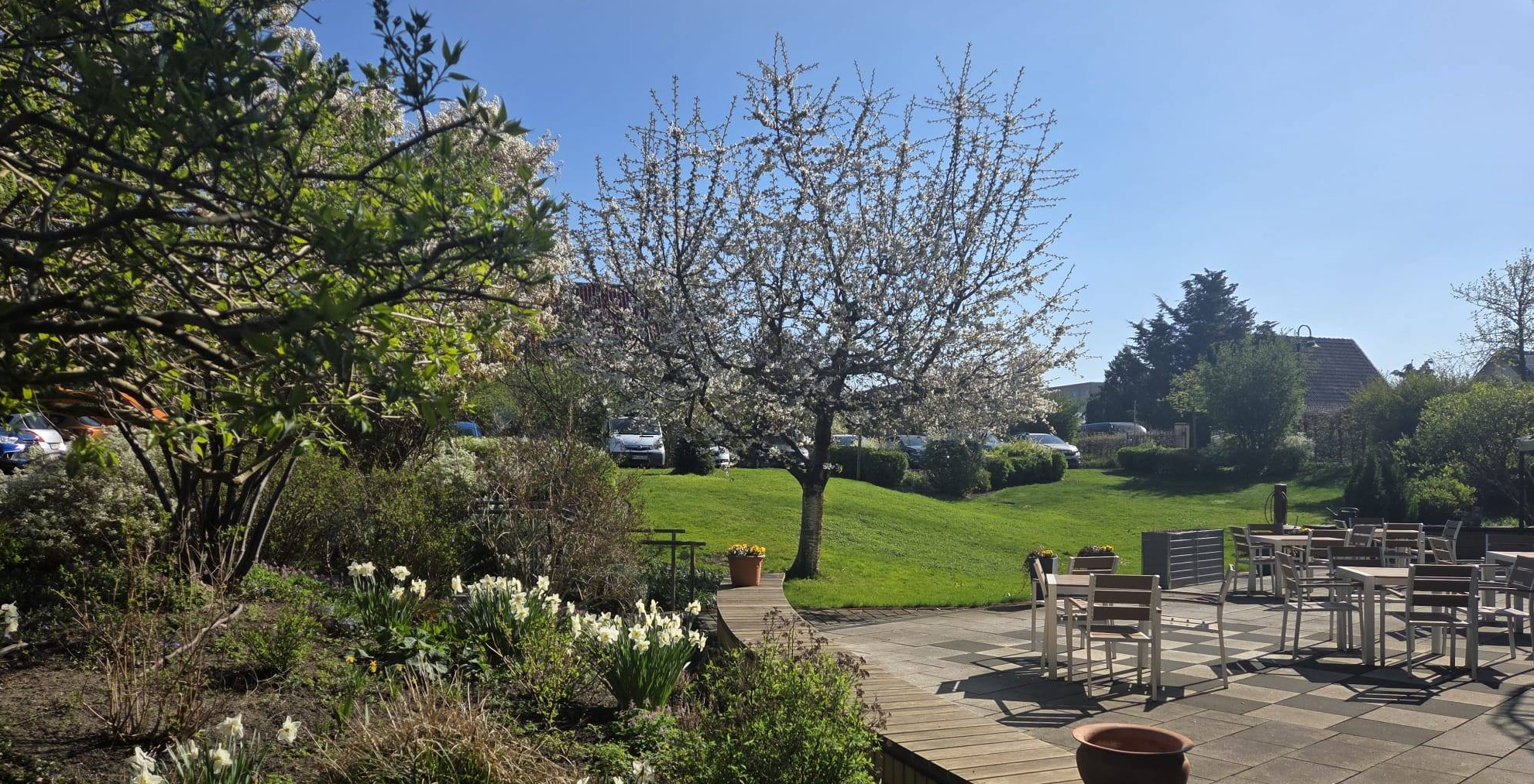Links: Blumen, Bäume, Sträucher. Rechts Terrasse mit Tischen und Stühlen in der Sonne.