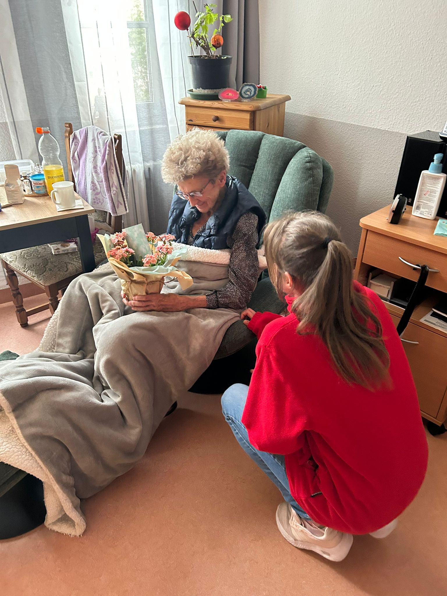 Bewohnerzimmer. Eine ältere Frau mit eingewickelten Blumentopf in den Händen sitzt in einem grünen Sessel mit eine grauen Decke auf den Beinen.