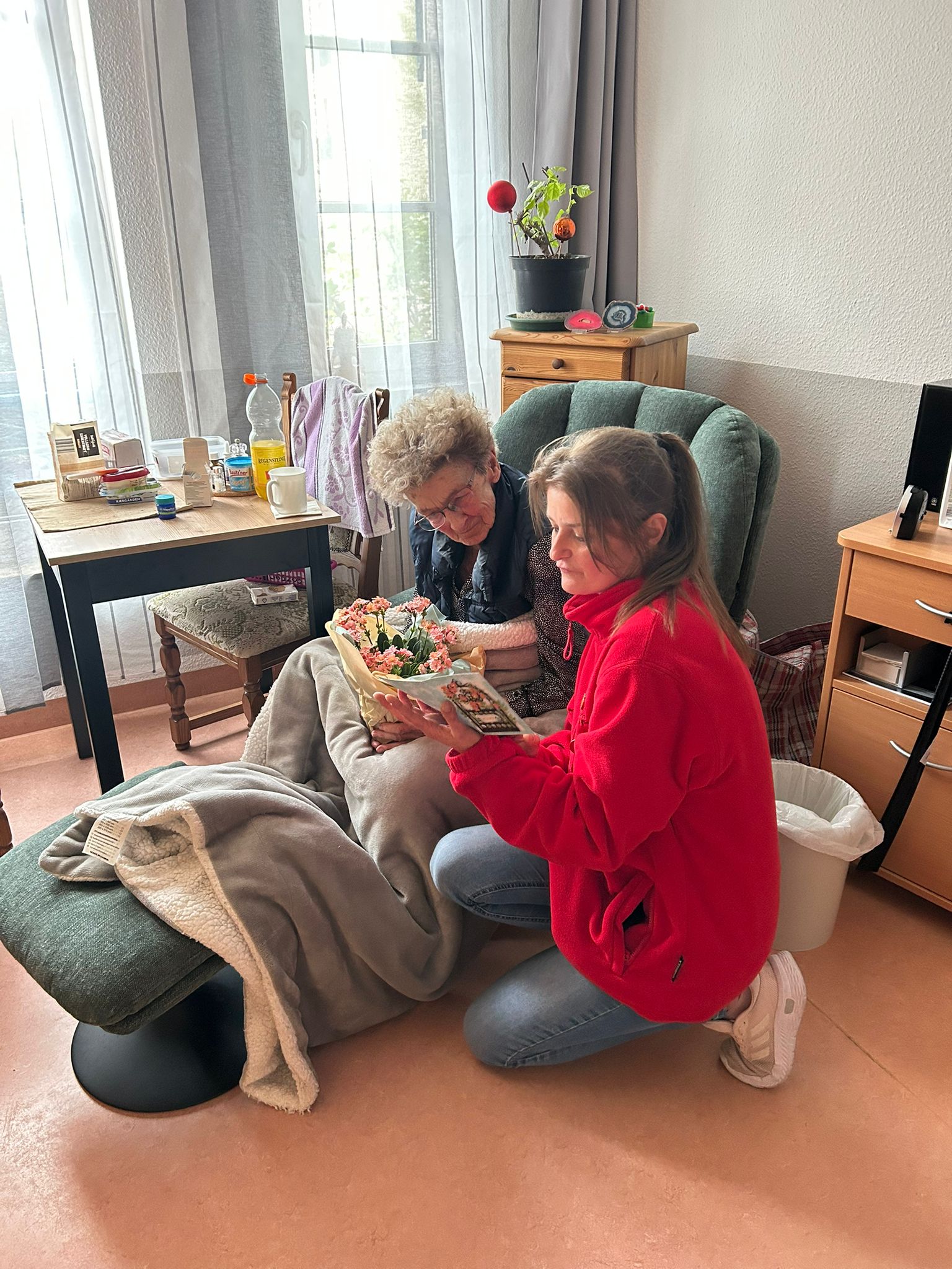 Bewohnerzimmer: Mitarbeiterin kniet mit Blumen und Karte neben einer Seniorin im grünen Sessel.