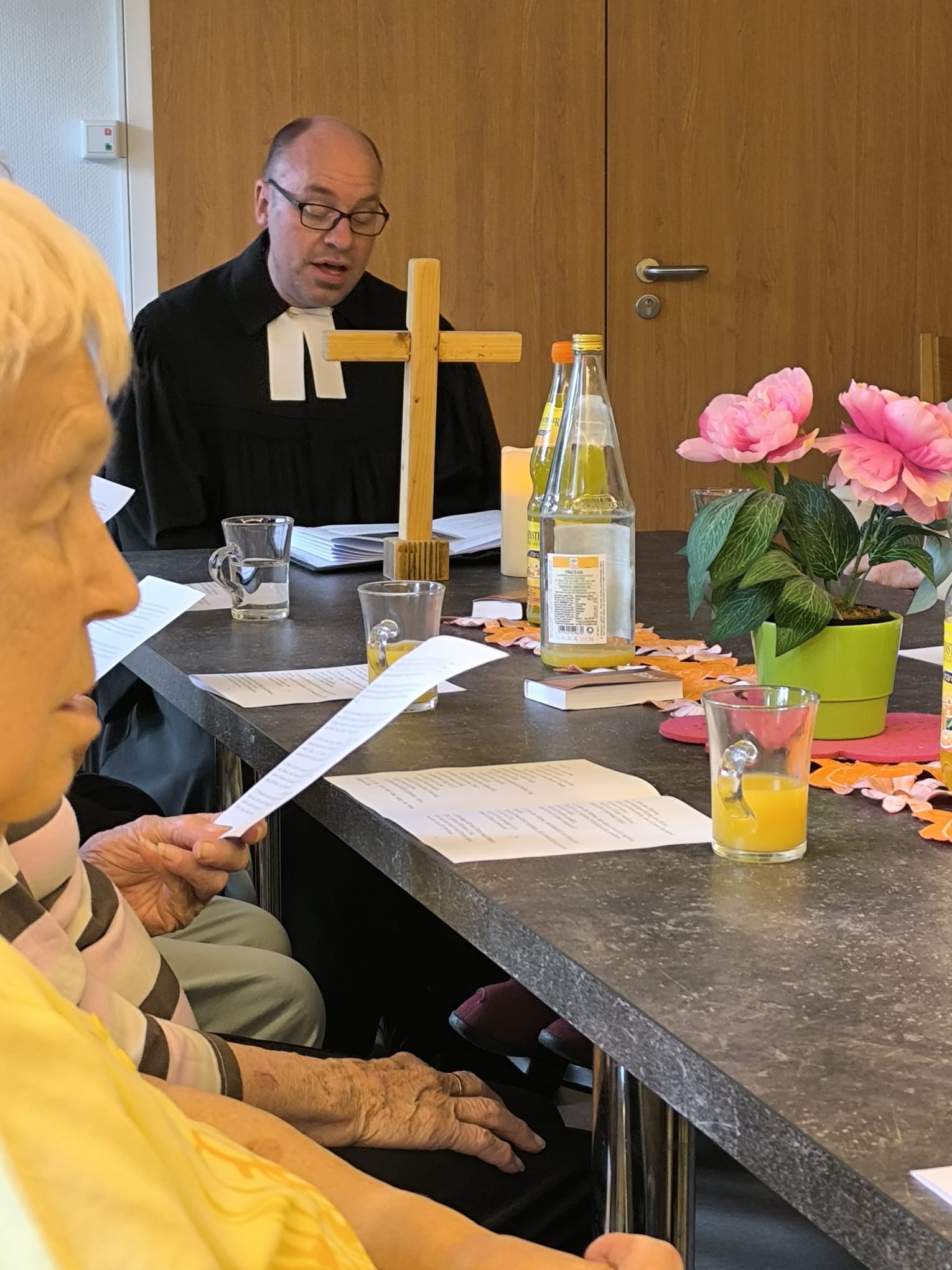 Ein Tisch. Am Kopfende sitzt der Pfarrer, ein Holzkreuz steht vor ihm. Am Tisch sitzen Senioren.